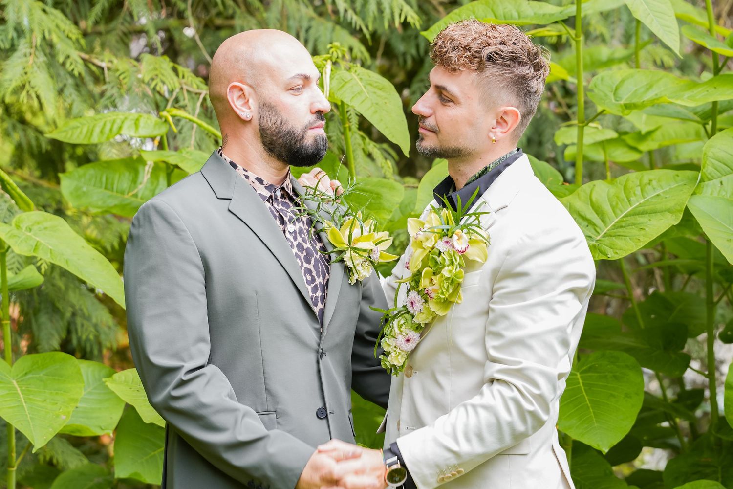 Two grooms in suits exchange vows outdoors surrounded by lush tropical greenery, wearing floral leis.