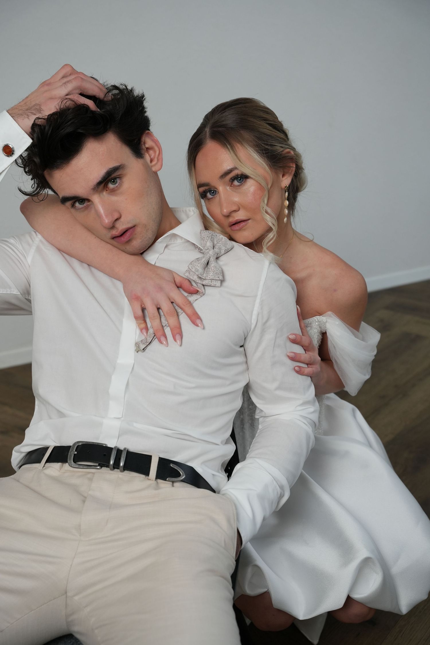 A stylish couple in white outfits pose on the floor, the woman embracing the man from behind in an elegant editorial shoot.