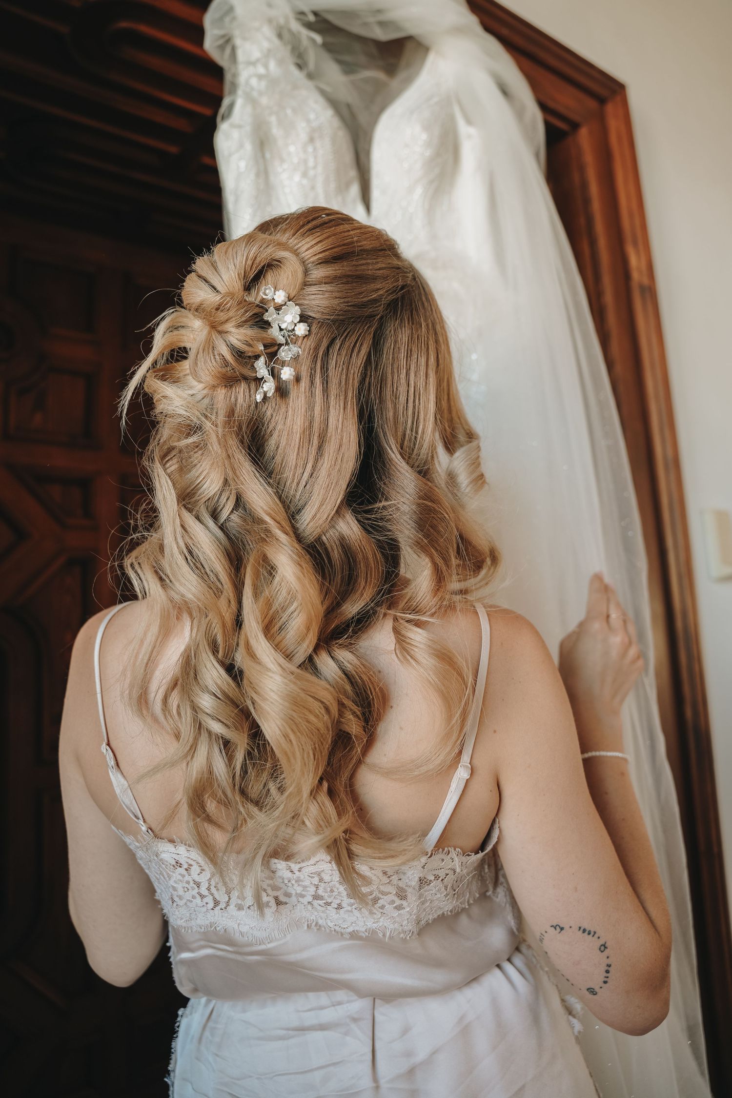 Bride with half-up curly hairstyle adorned with pearl pins, wearing a lace slip dress before her wedding gown.