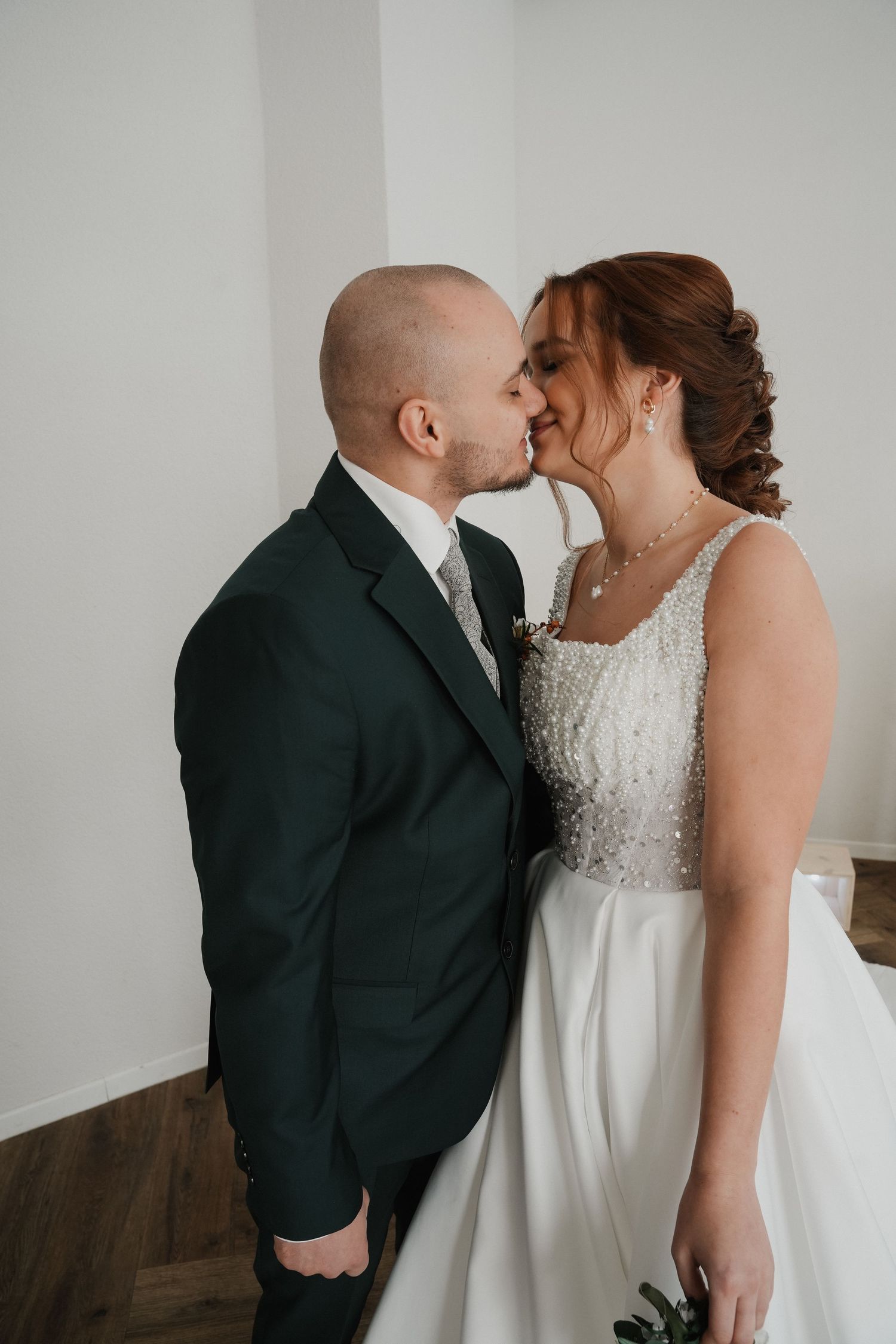 A bride and groom share a romantic kiss in a bright white room, she in a beaded gown, he in a dark green suit.