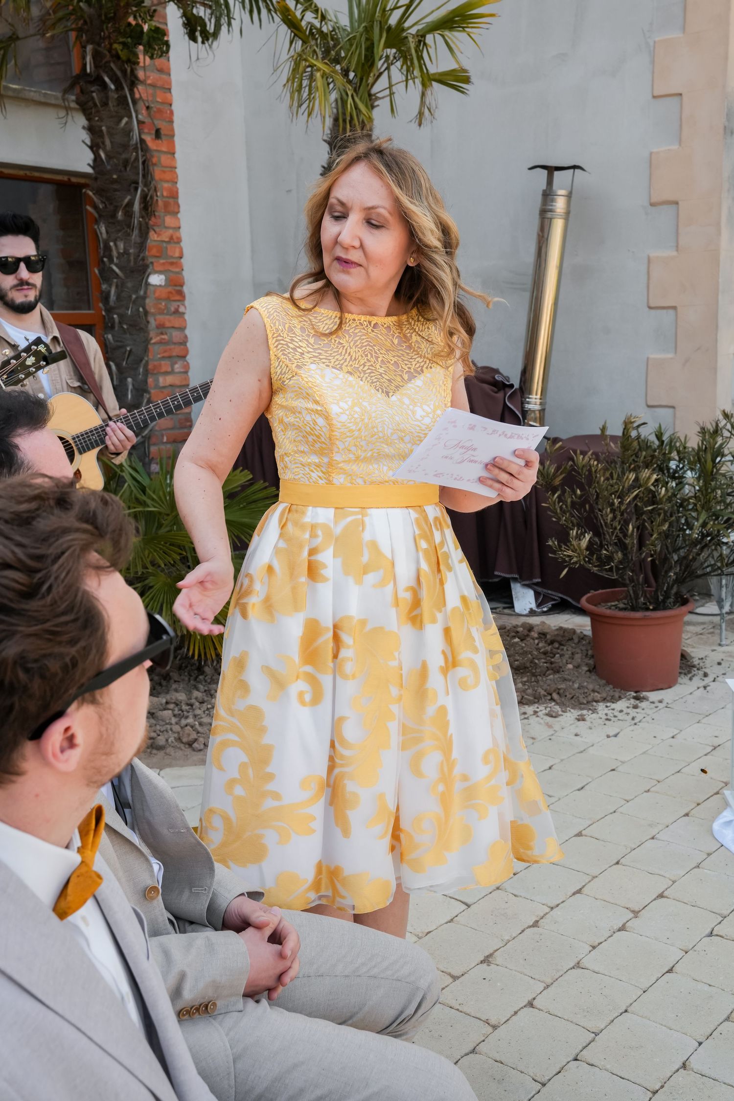 Woman in yellow floral dress reads at outdoor wedding ceremony while guitarist performs in background.