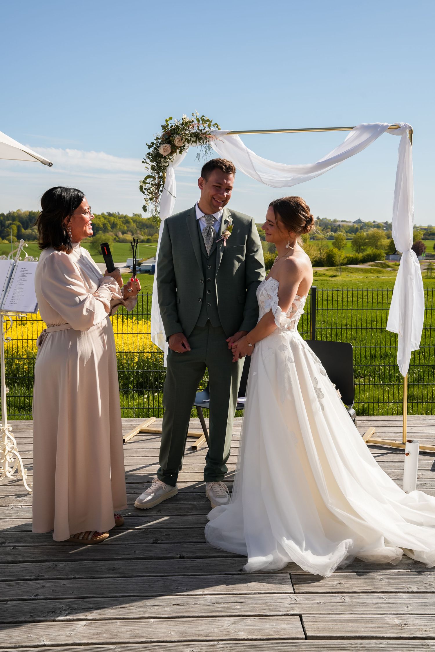 Outdoor wedding ceremony with bride in white gown, groom in gray suit, and officiant on a wooden deck.
