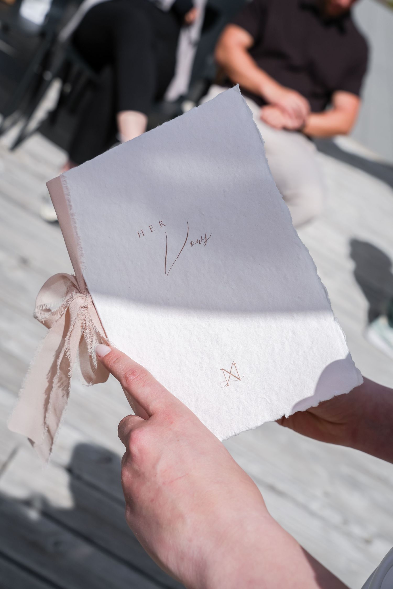 Hands holding a elegant white wedding vow booklet tied with ribbon, labeled Her Vows, at an outdoor ceremony.
