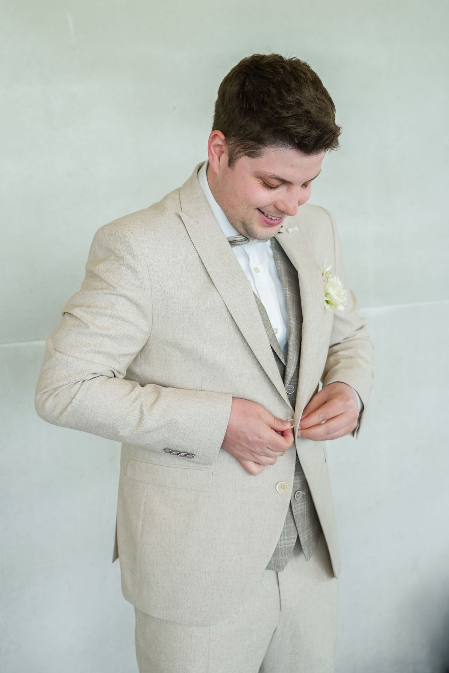 Smiling groom in beige linen suit with boutonniere buttoning jacket before wedding ceremony.