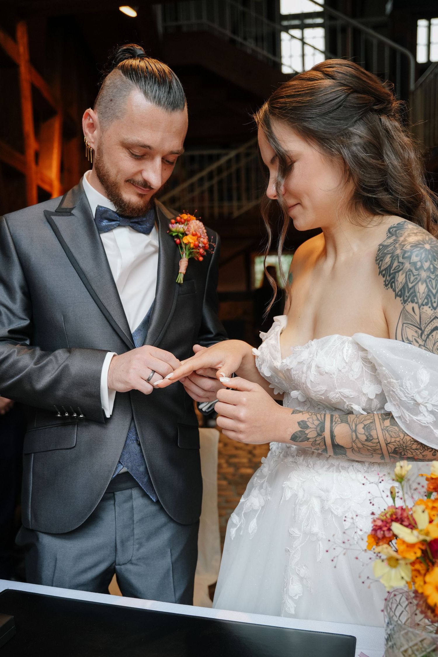Bride with floral tattoos and groom in a gray suit exchange rings during their wedding ceremony, with colorful flower bouquet nearby.