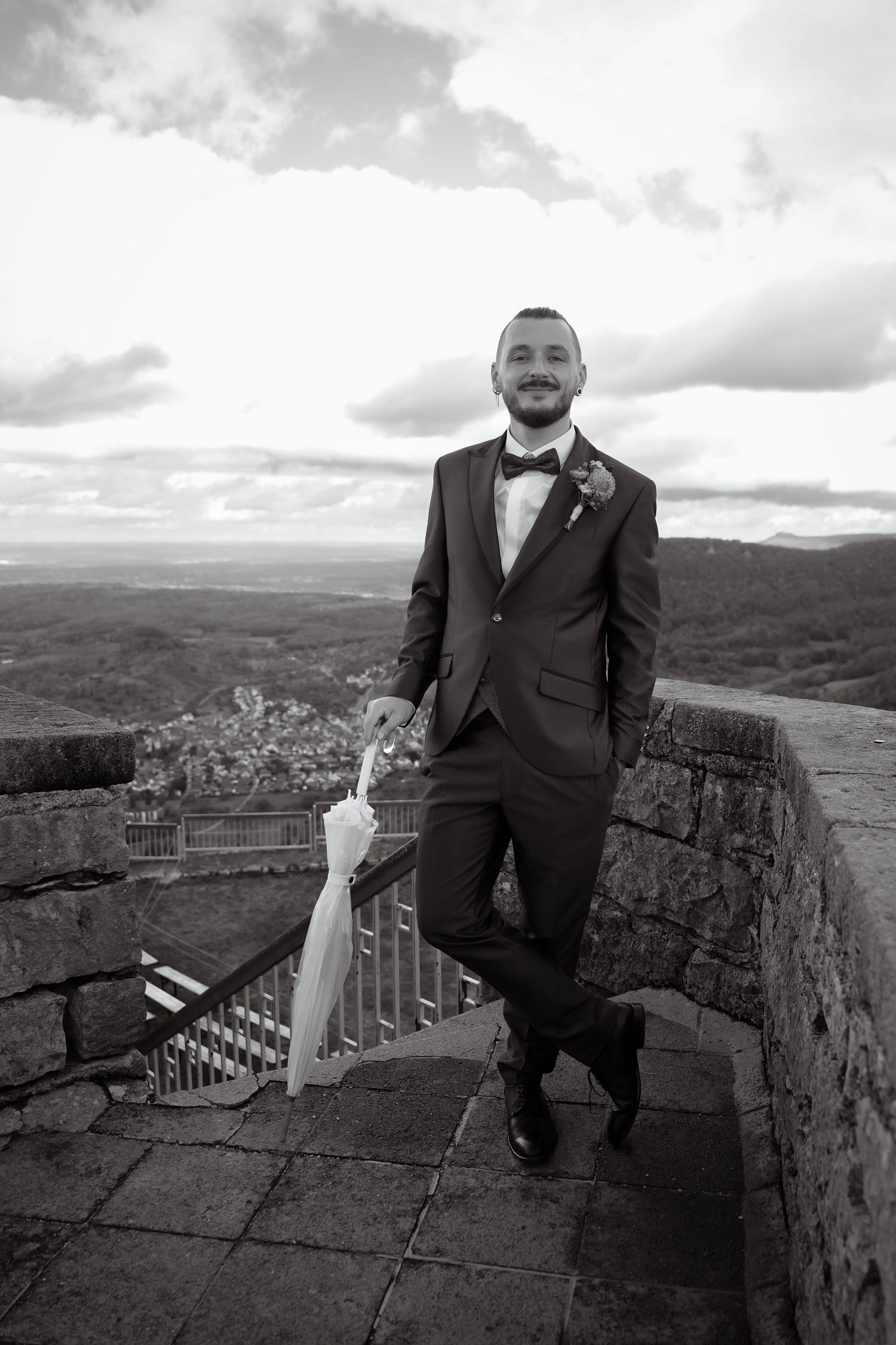 Black and white photo of a groom in a suit and bow tie standing on a stone terrace with a folded umbrella and a scenic view.