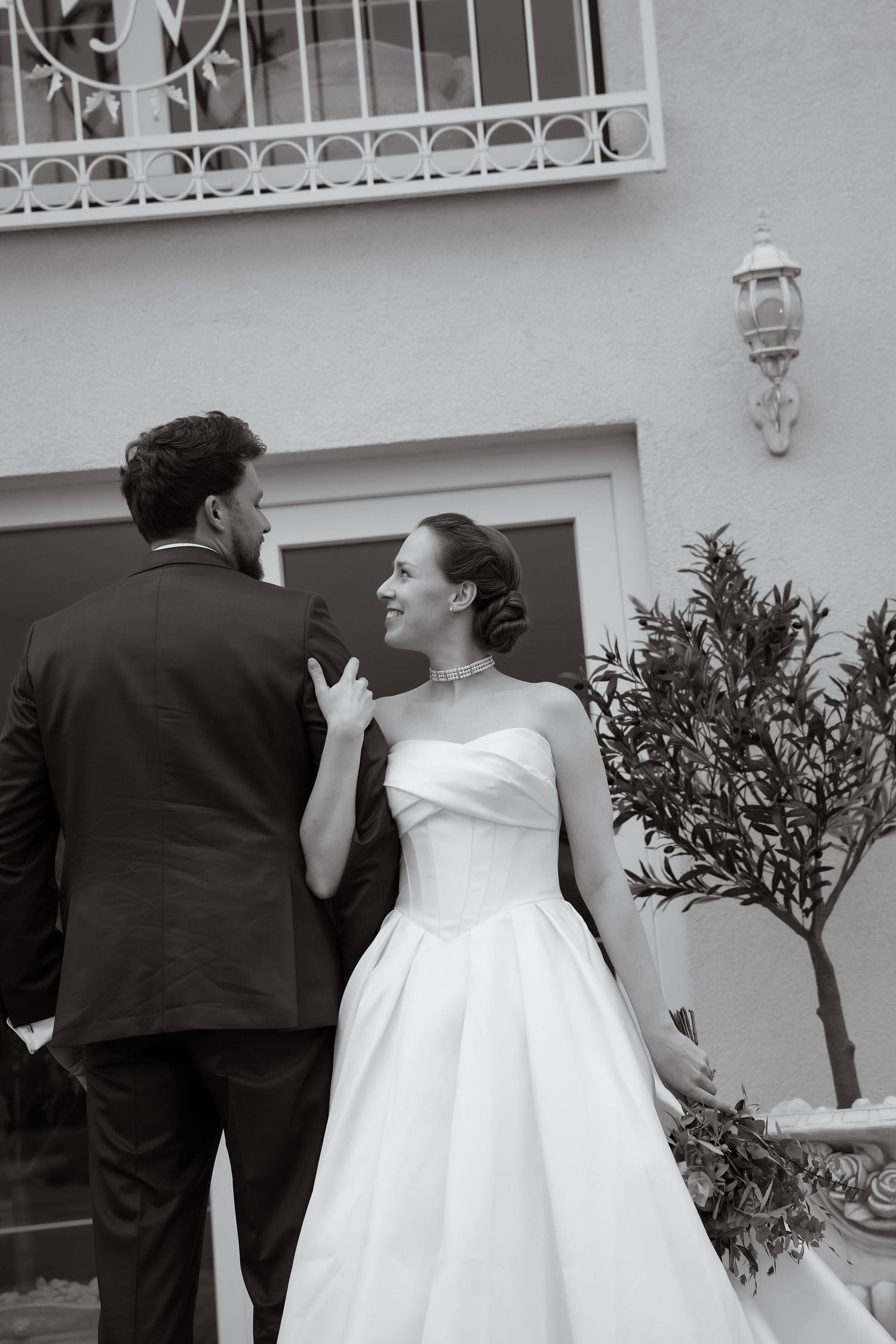 Black and white wedding photo of a bride in a strapless gown smiling up at her groom in a dark suit beside an olive tree.
