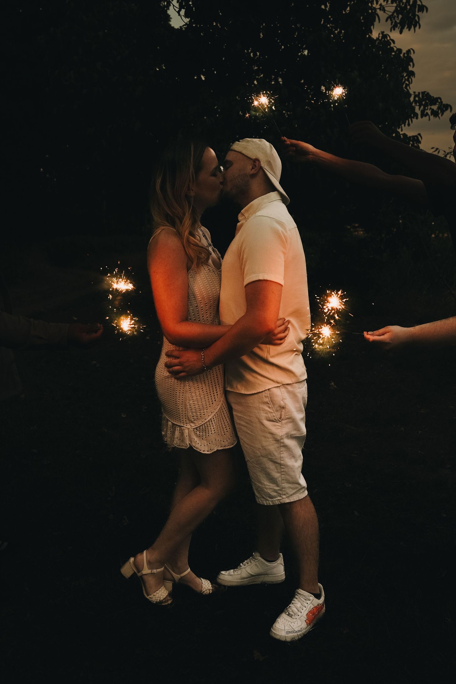 Couple kissing outdoors at night surrounded by glowing sparklers held by friends in a romantic celebration.