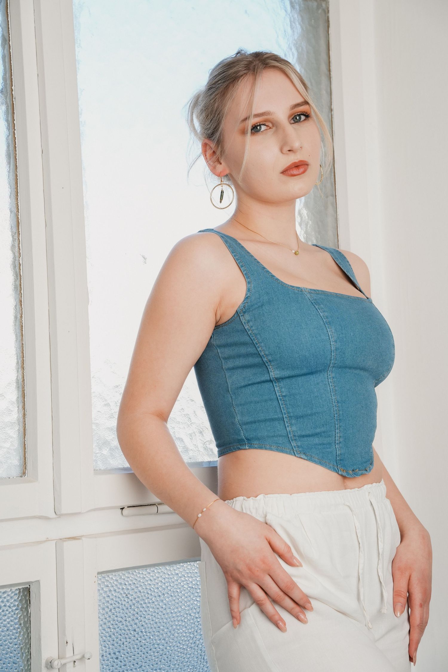Young blonde woman in denim crop top and white pants posing near a window with natural light.