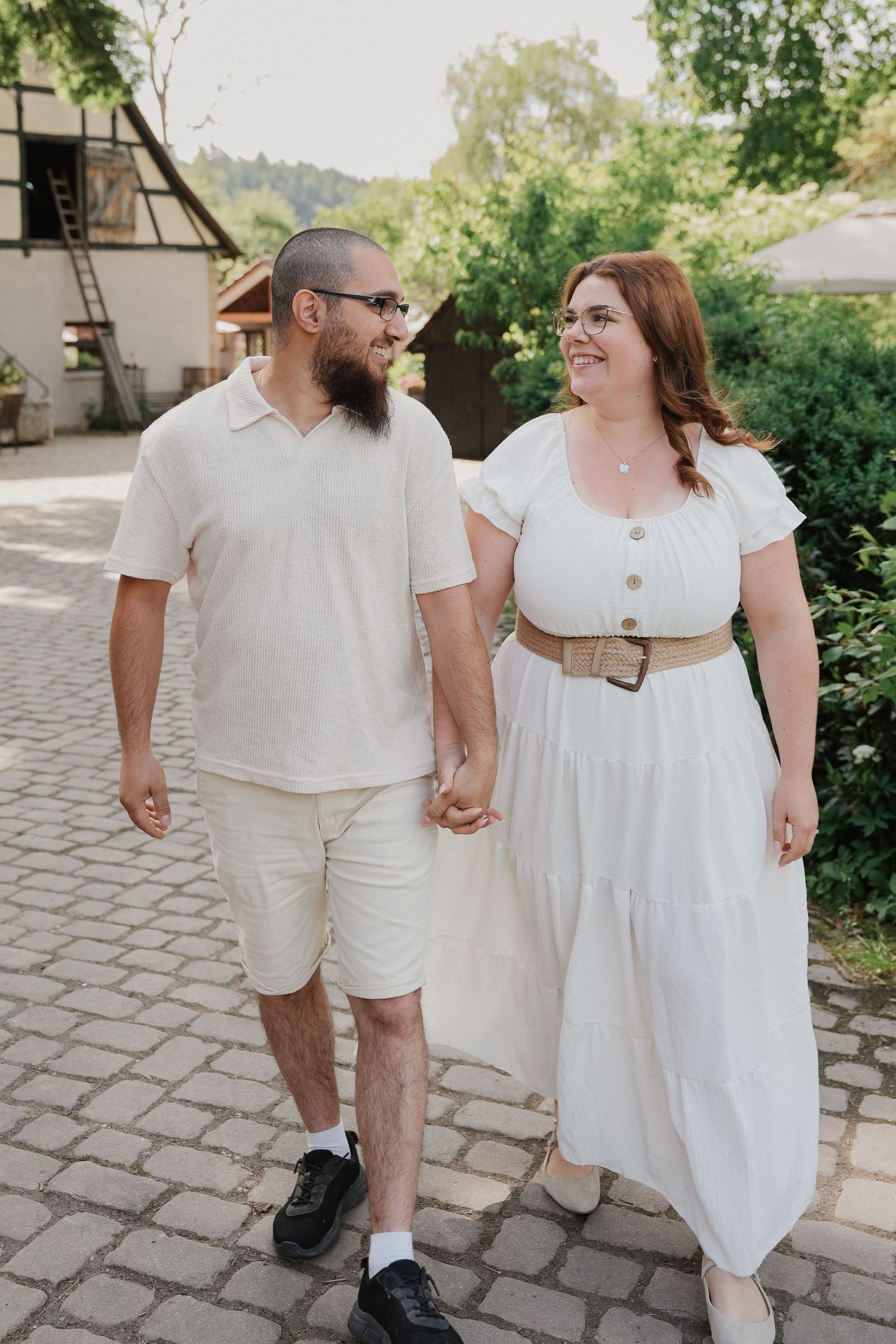 A couple in white outfits holds hands while walking on cobblestone, smiling at each other outdoors.