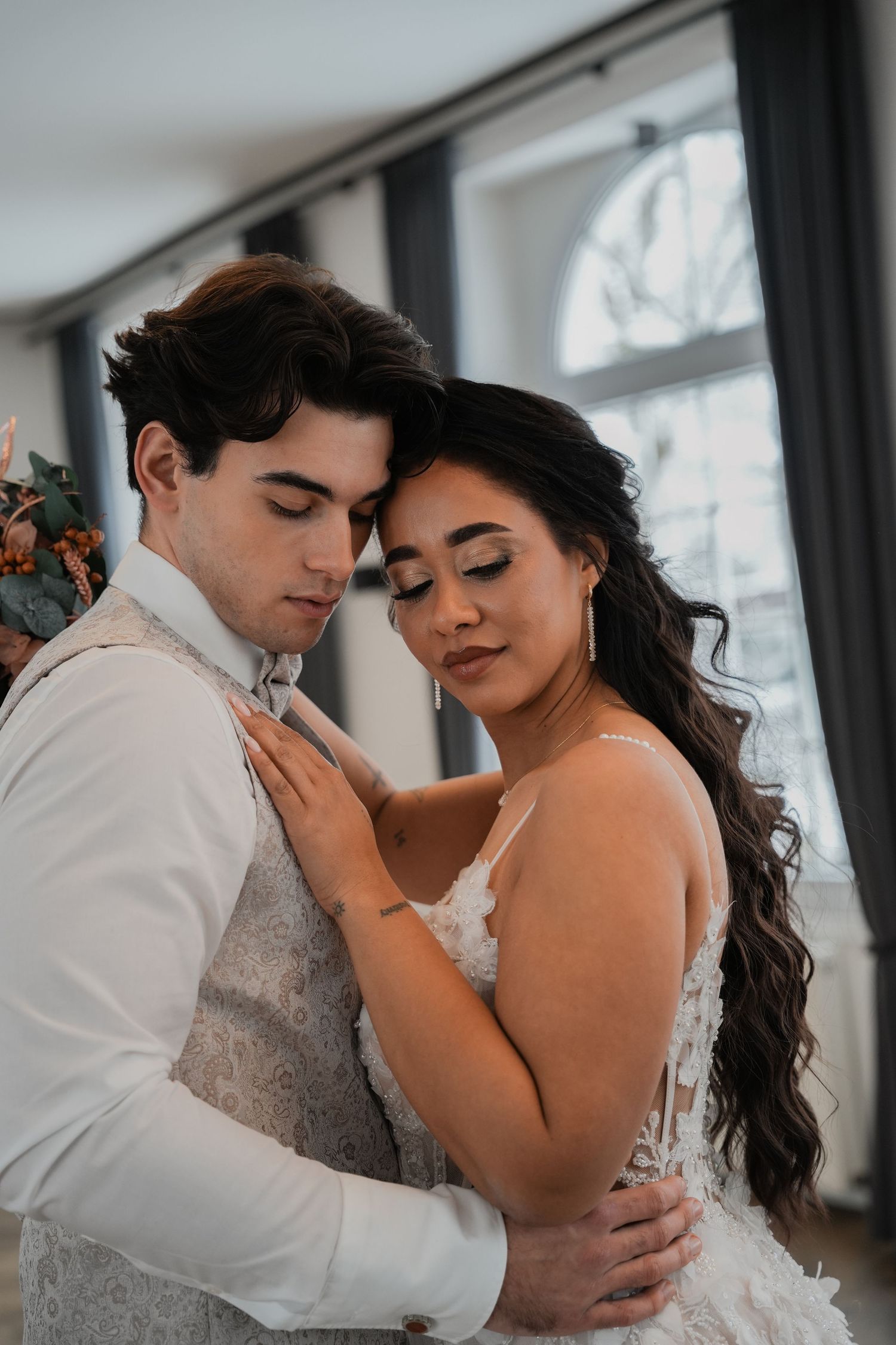 A romantic moment between a couple in wedding attire near a bright window.