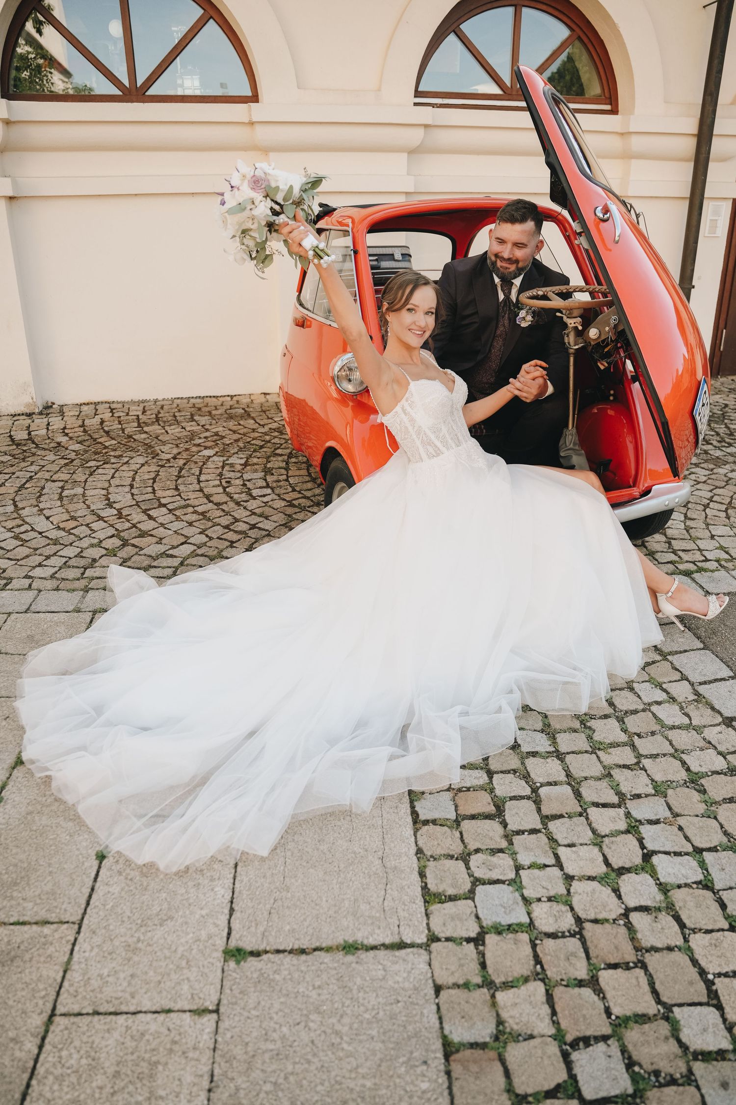 Bride in white gown holds bouquet beside groom exiting a vintage red microcar on cobblestone.