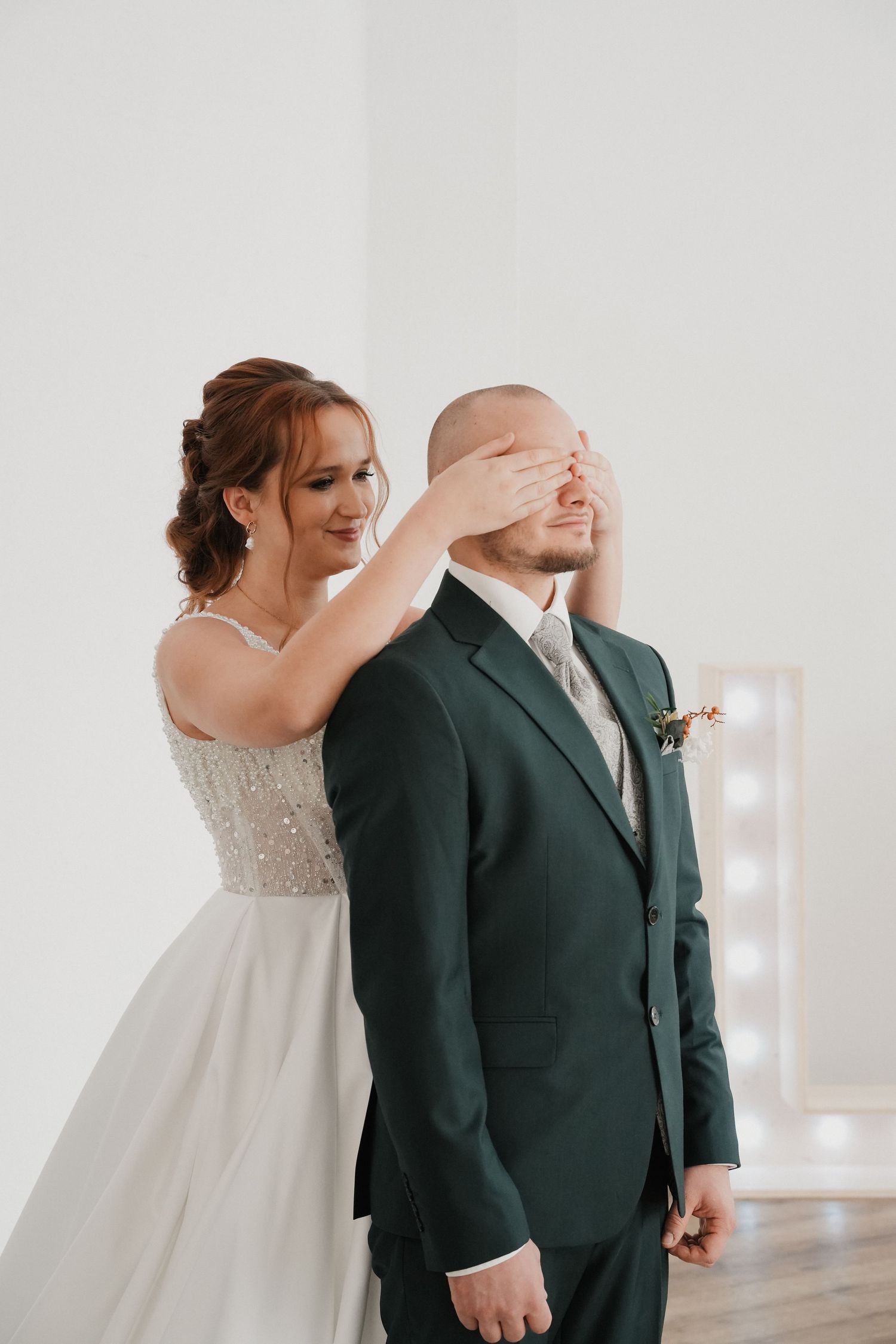 Bride covers groom's eyes from behind in a first look moment, wearing white gown as he stands in a green suit.