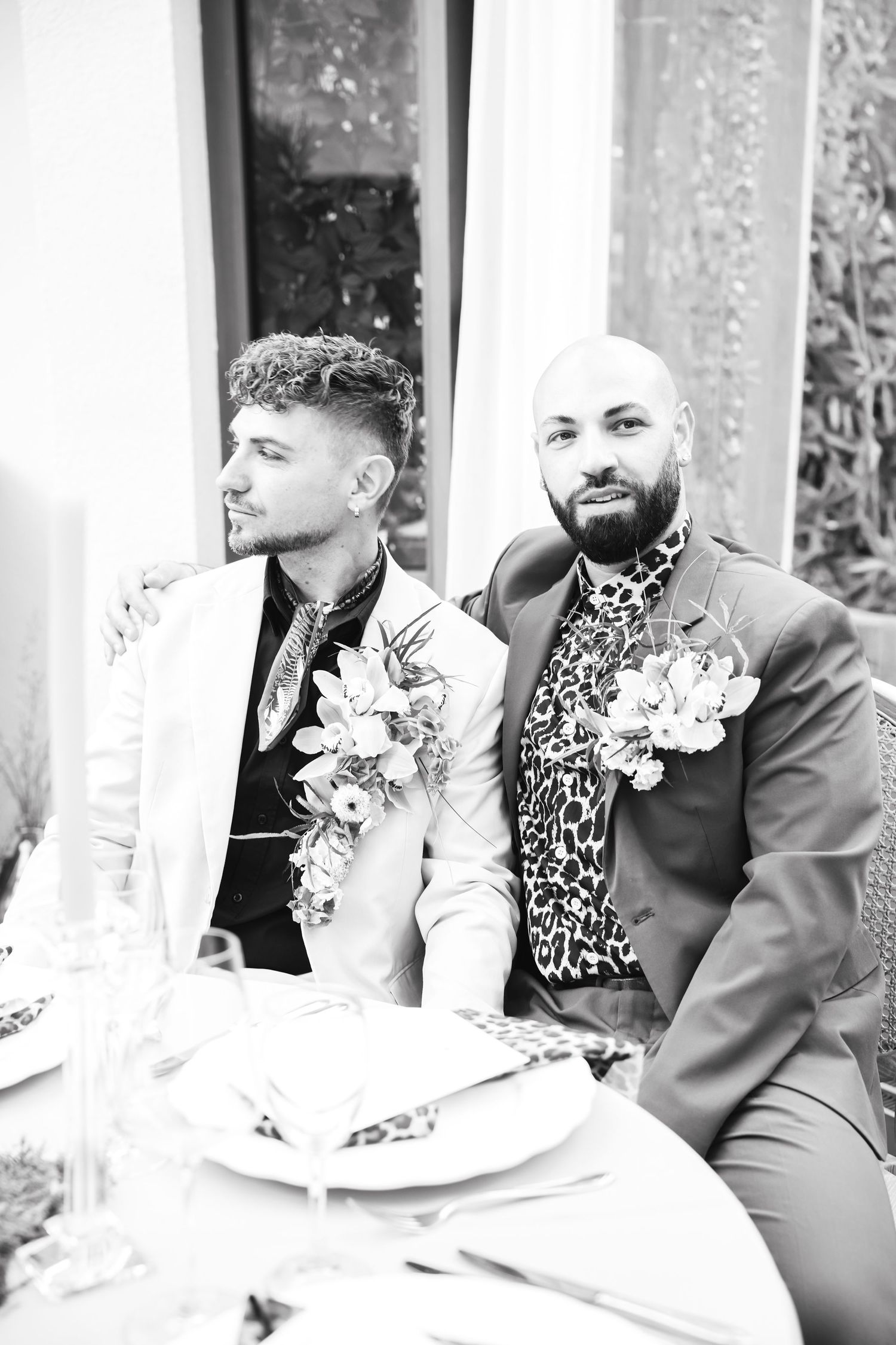 Black and white photo of two grooms in floral boutonnieres sitting together at wedding reception table.