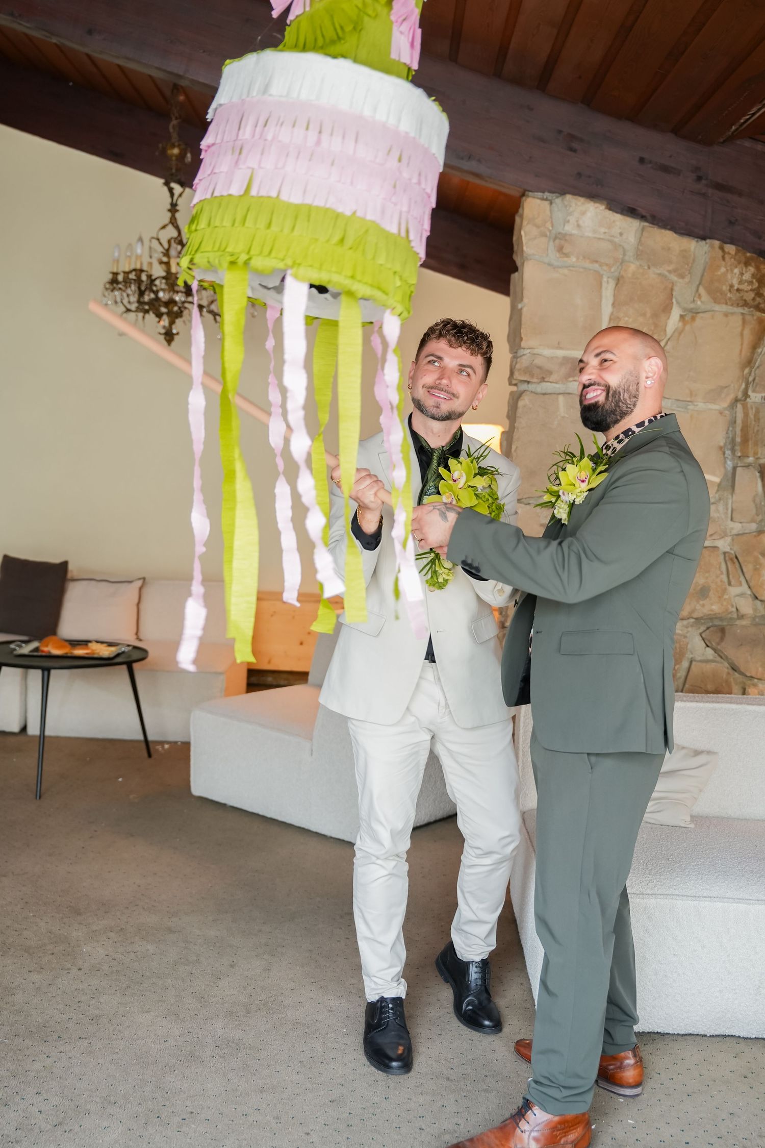 Two grooms in suits smile while holding a colorful cake-shaped piñata at their wedding celebration indoors.