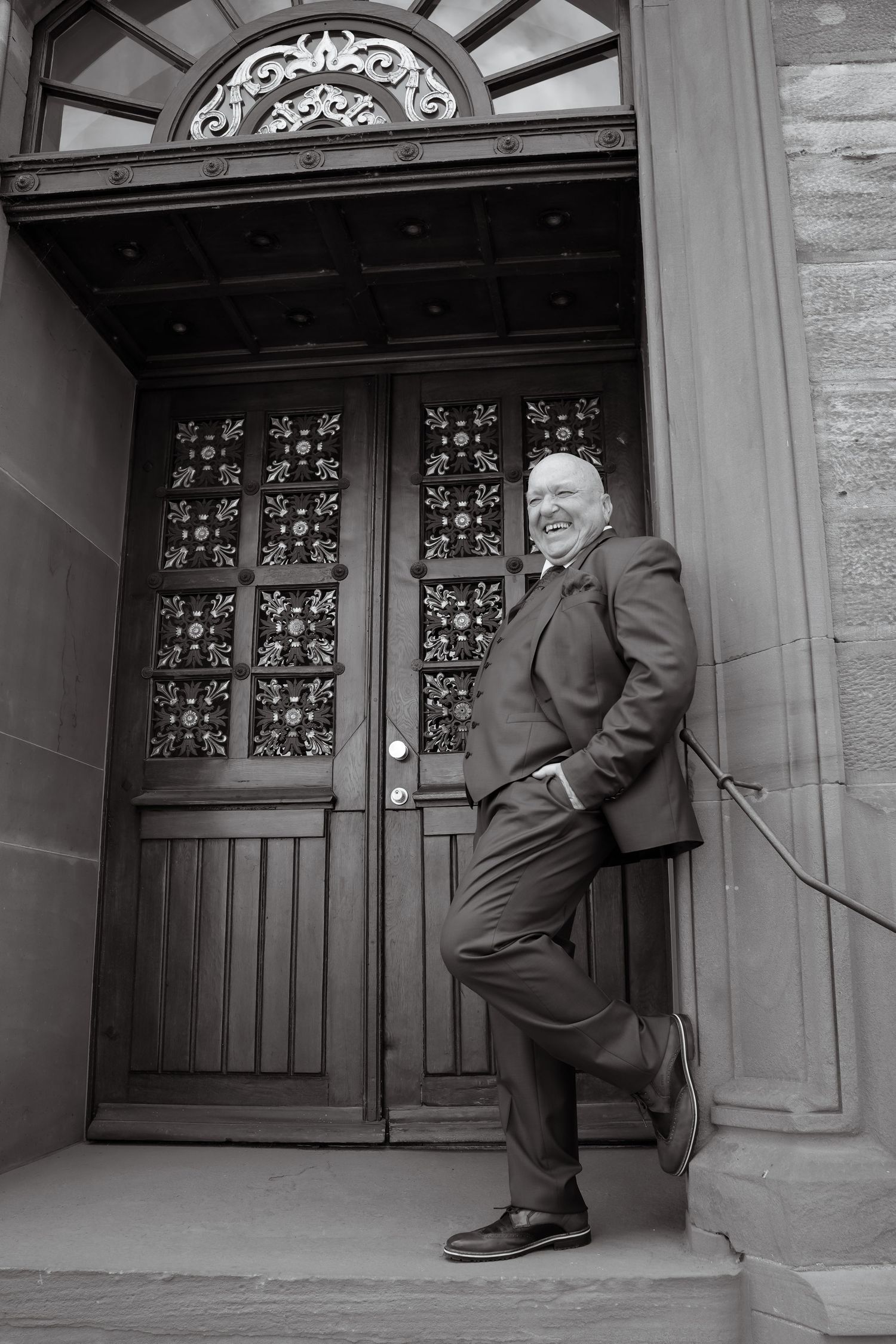 Man in suit smiling and leaning against ornate wooden doors in black and white portrait photo.