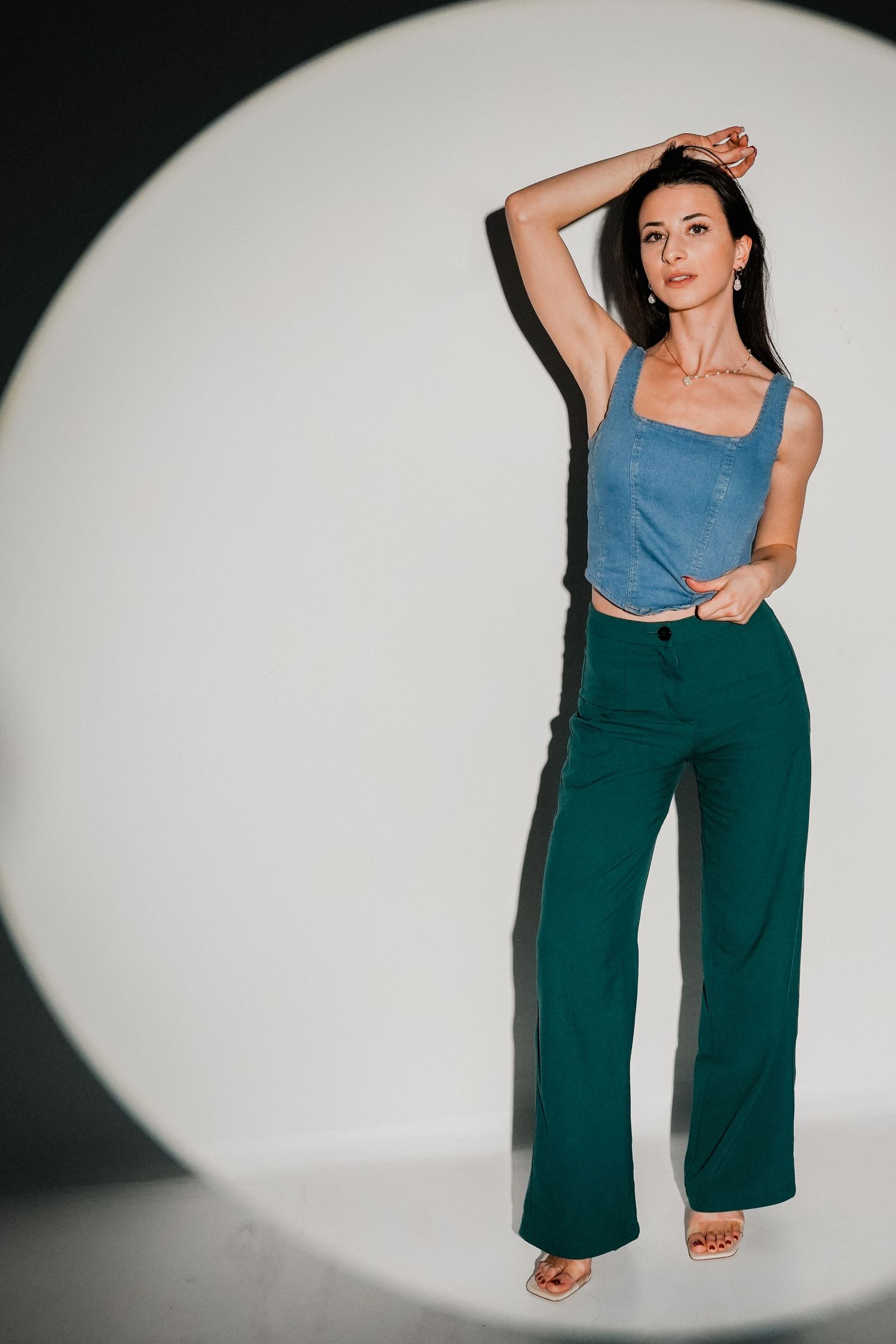 Woman posing in blue crop top and green wide-leg trousers against white circular backdrop in studio.