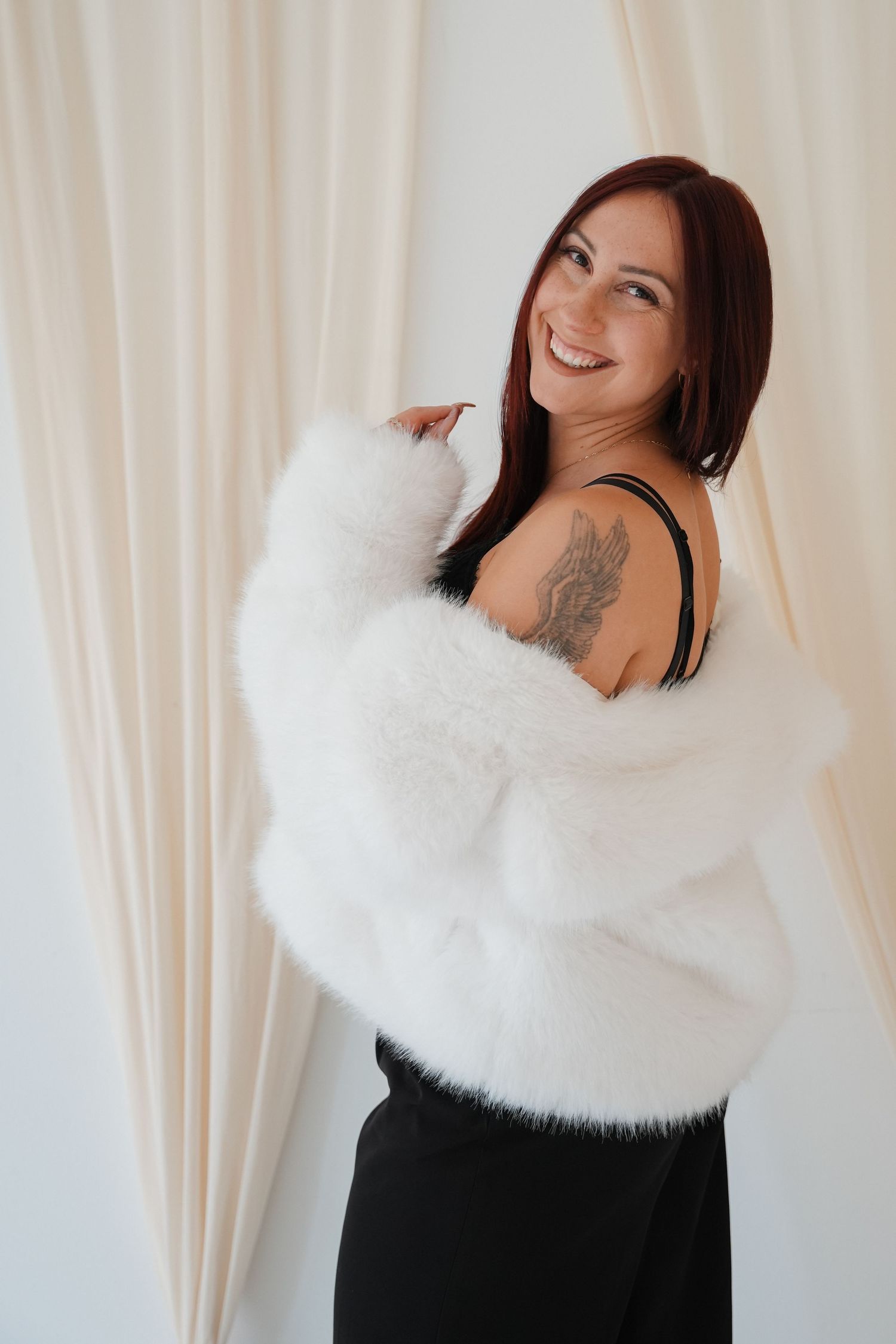 Smiling woman with red hair holds a fluffy white cat, wearing a black outfit with a shoulder tattoo, against a light curtain.