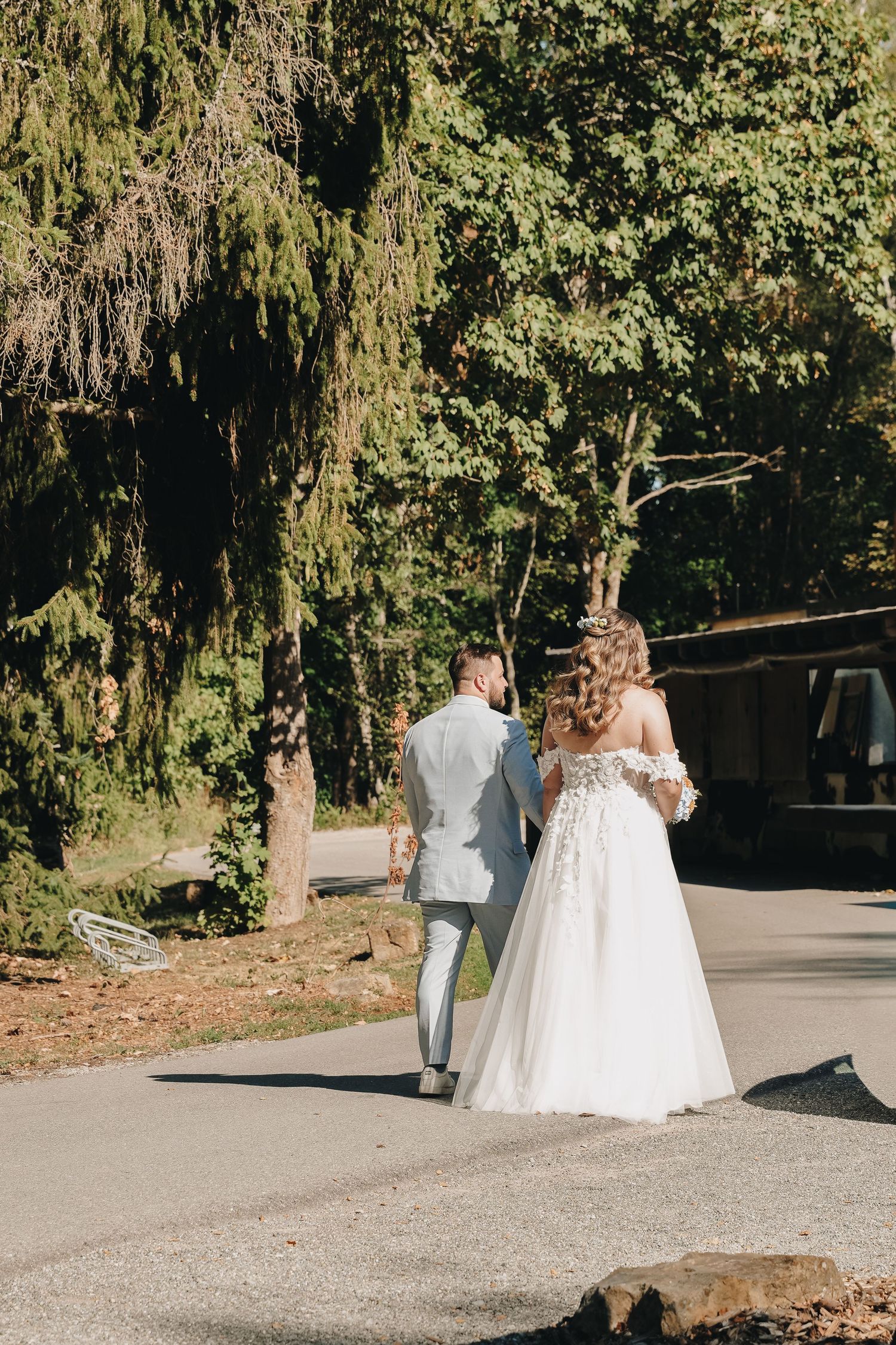 Bride and groom walking hand in hand through a sunlit park, viewed from behind, surrounded by lush green trees.