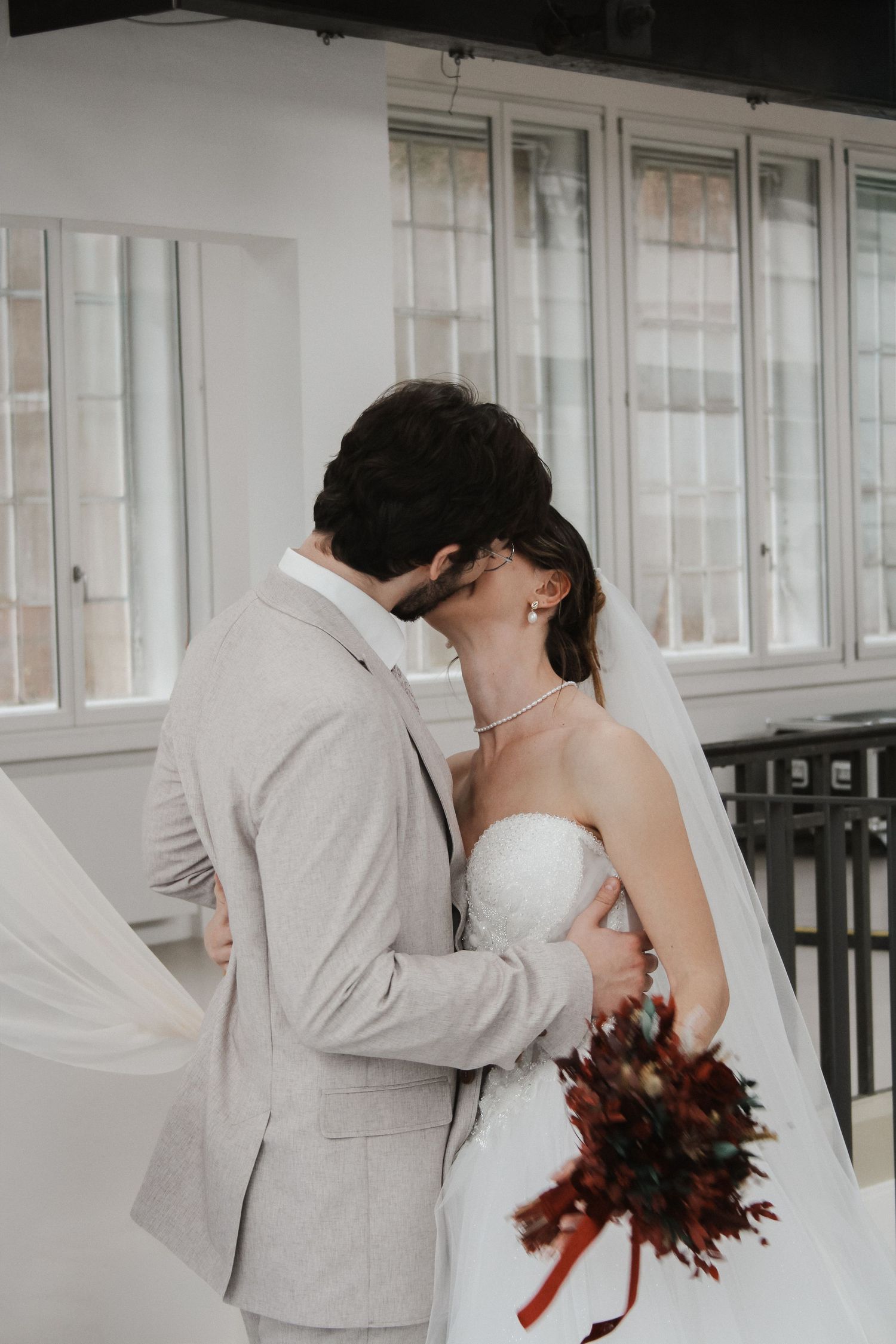 Bride and groom sharing a kiss indoors, bride in white strapless gown with veil holding red bouquet, groom in light gray suit.