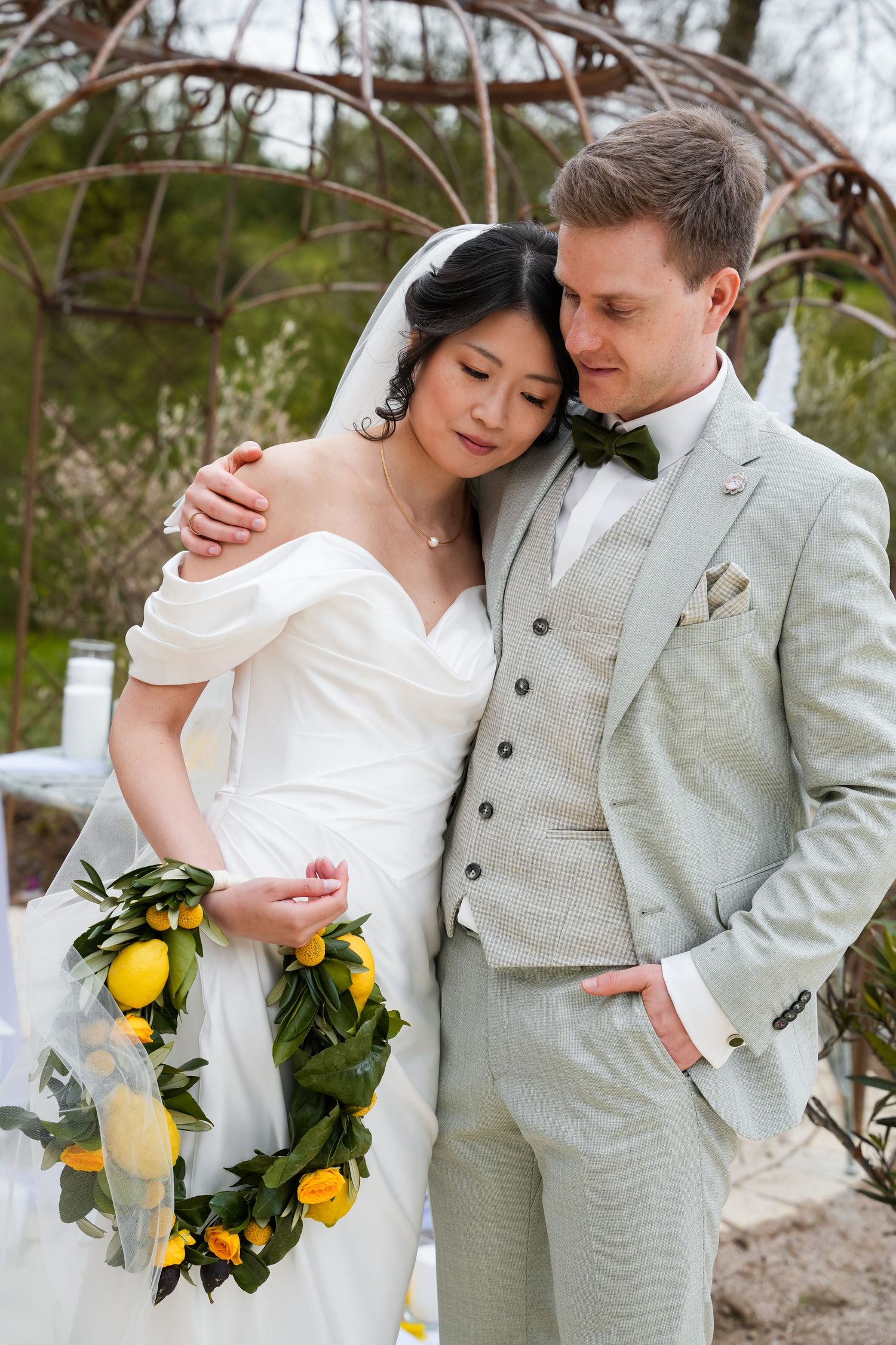 Bride and groom embrace outdoors, she holds a lemon wreath, wearing white gown, he wears a light gray suit.