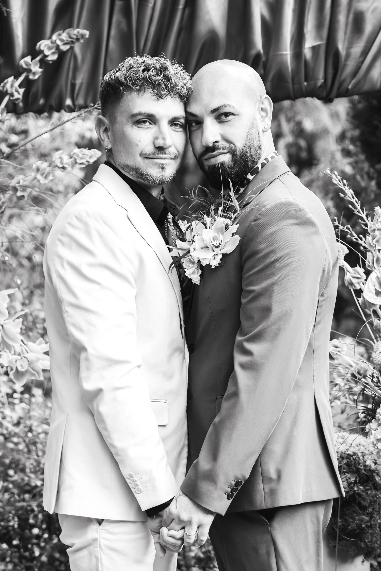 Black and white photo of two grooms in suits holding hands at their wedding ceremony outdoors.