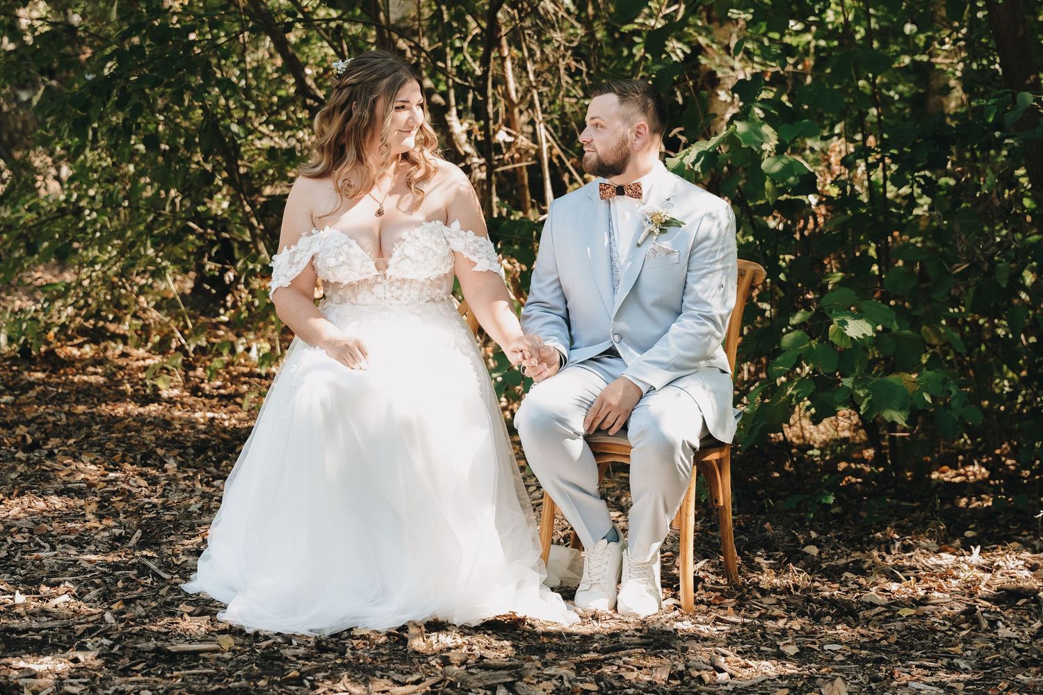 Bride and groom sitting on a wooden bench outdoors, surrounded by lush greenery, in wedding attire.