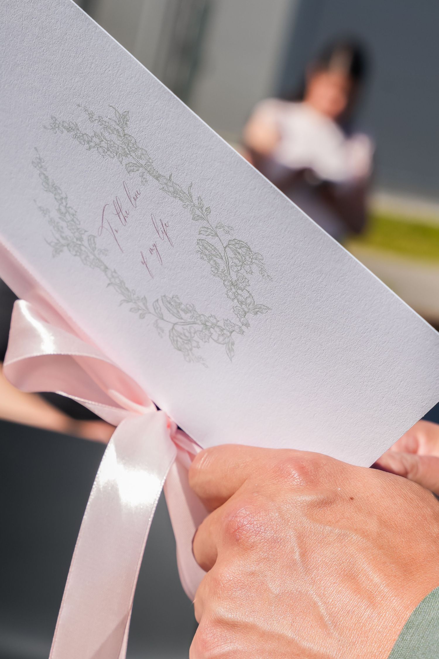 Hands holding an elegant white wedding program with pink ribbon bow and decorative floral border design.