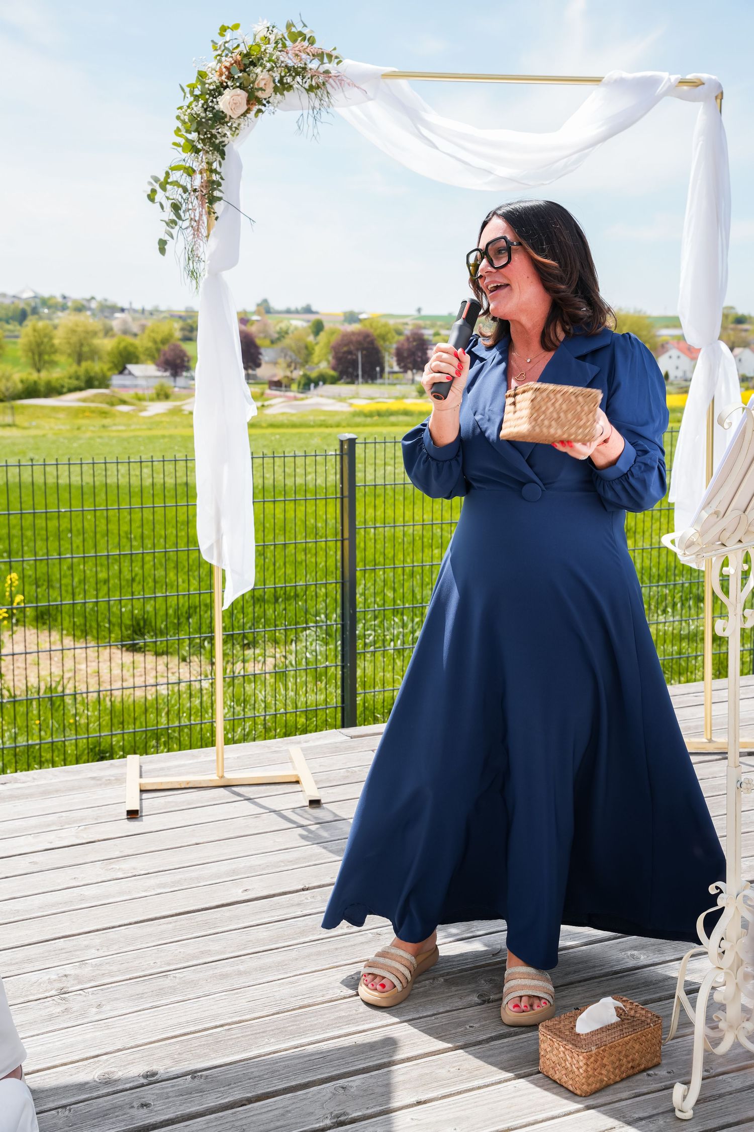 Woman in navy blue dress speaking at outdoor wedding ceremony near floral arch on sunny day.