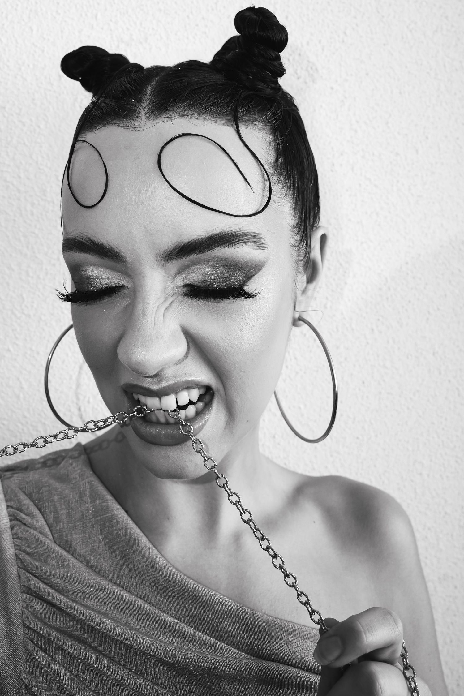 Black and white photo of a woman with space buns, hoop earrings, dramatic makeup, and a chain held in her teeth.