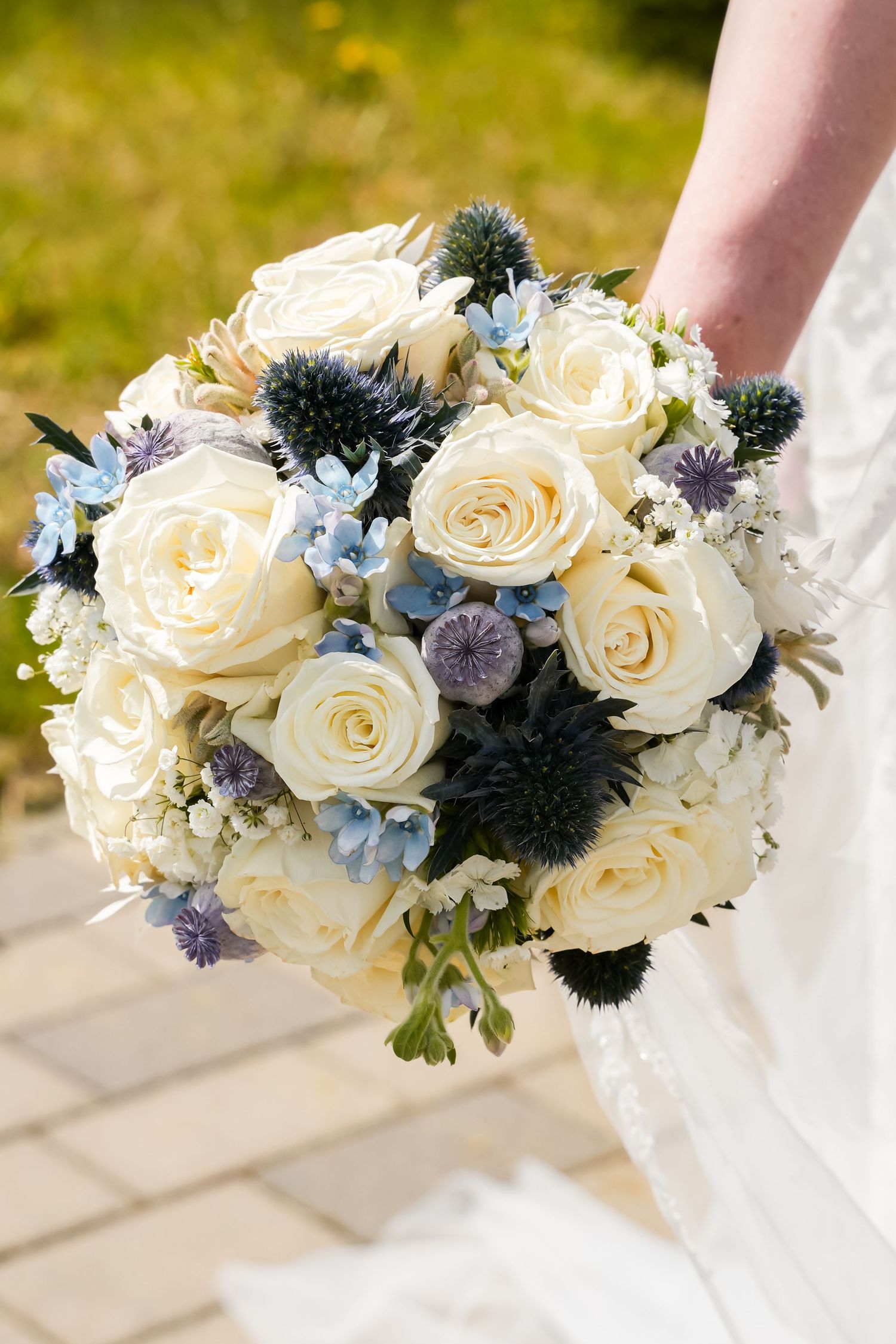 Bride holding a wedding bouquet of white roses, blue hydrangeas, and thistle flowers outdoors.