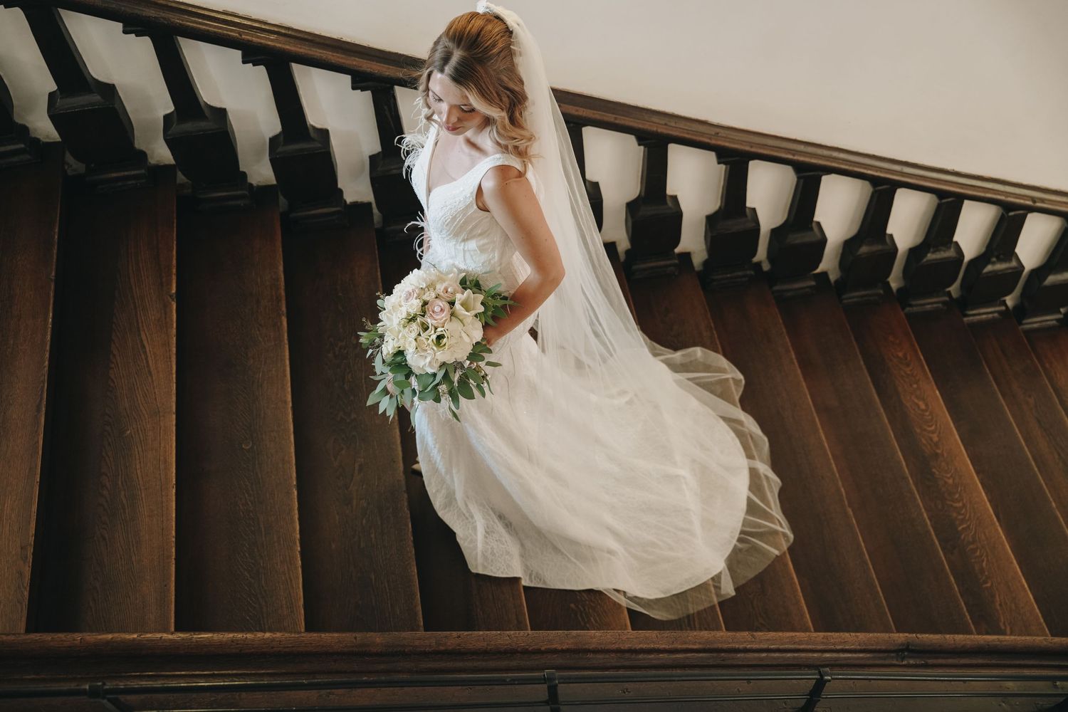 Bride in white wedding gown and veil holding bouquet descending elegant wooden staircase.