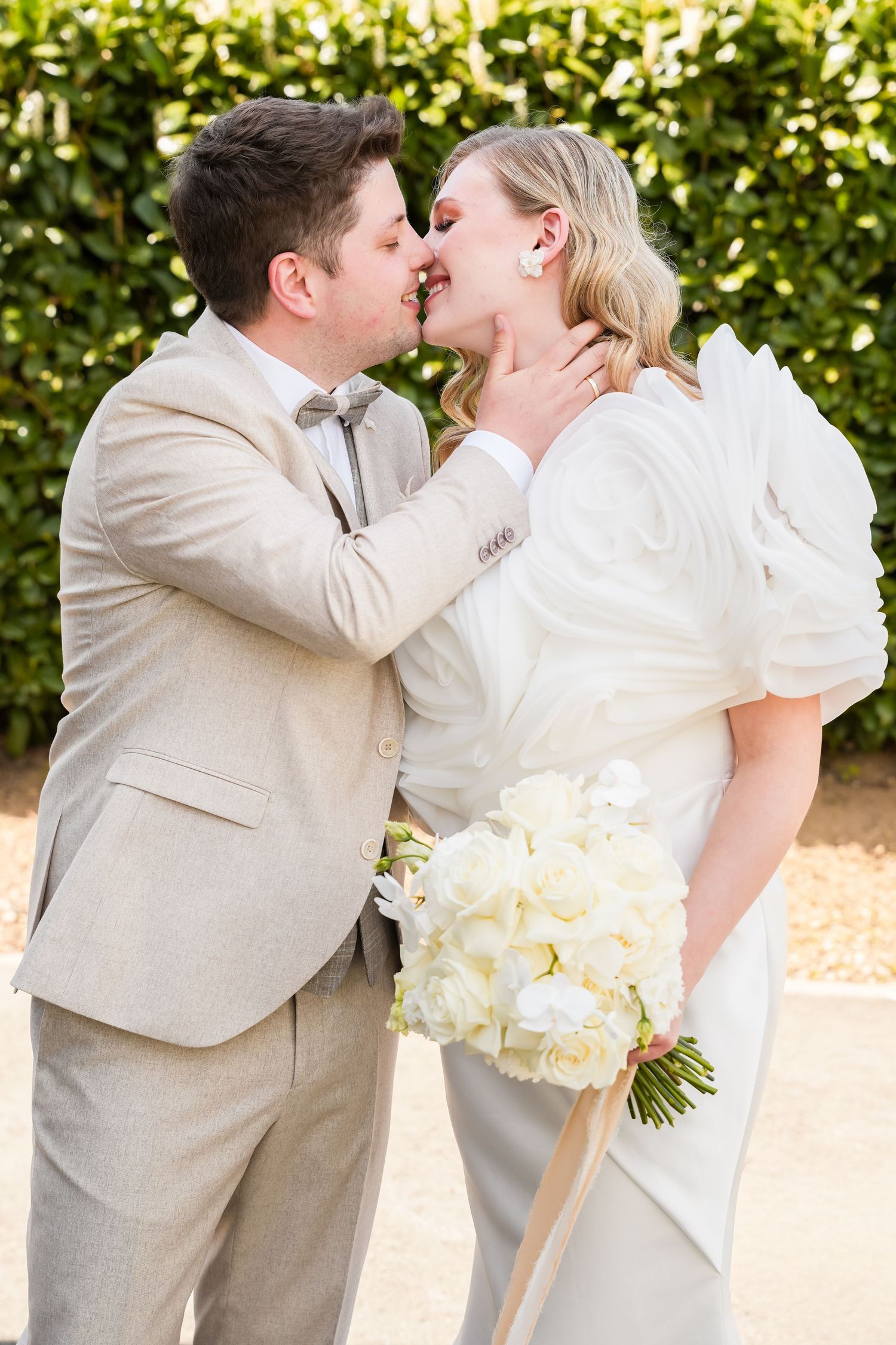 A bride and groom share a romantic kiss outdoors, she holds a white bouquet, wearing a white gown and he a beige suit.
