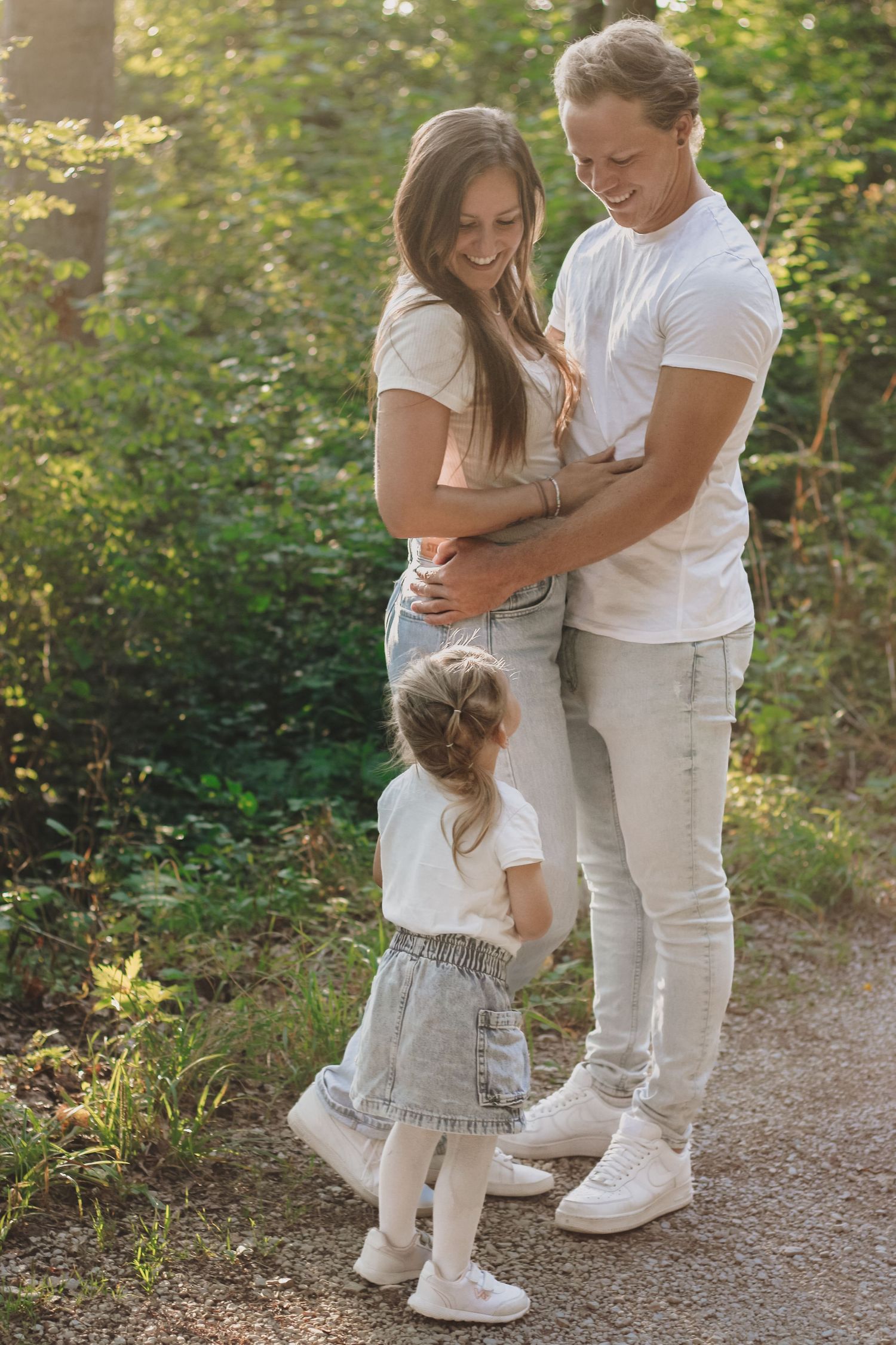 Happy young family with toddler daughter embracing on a sunny forest path in casual white outfits.