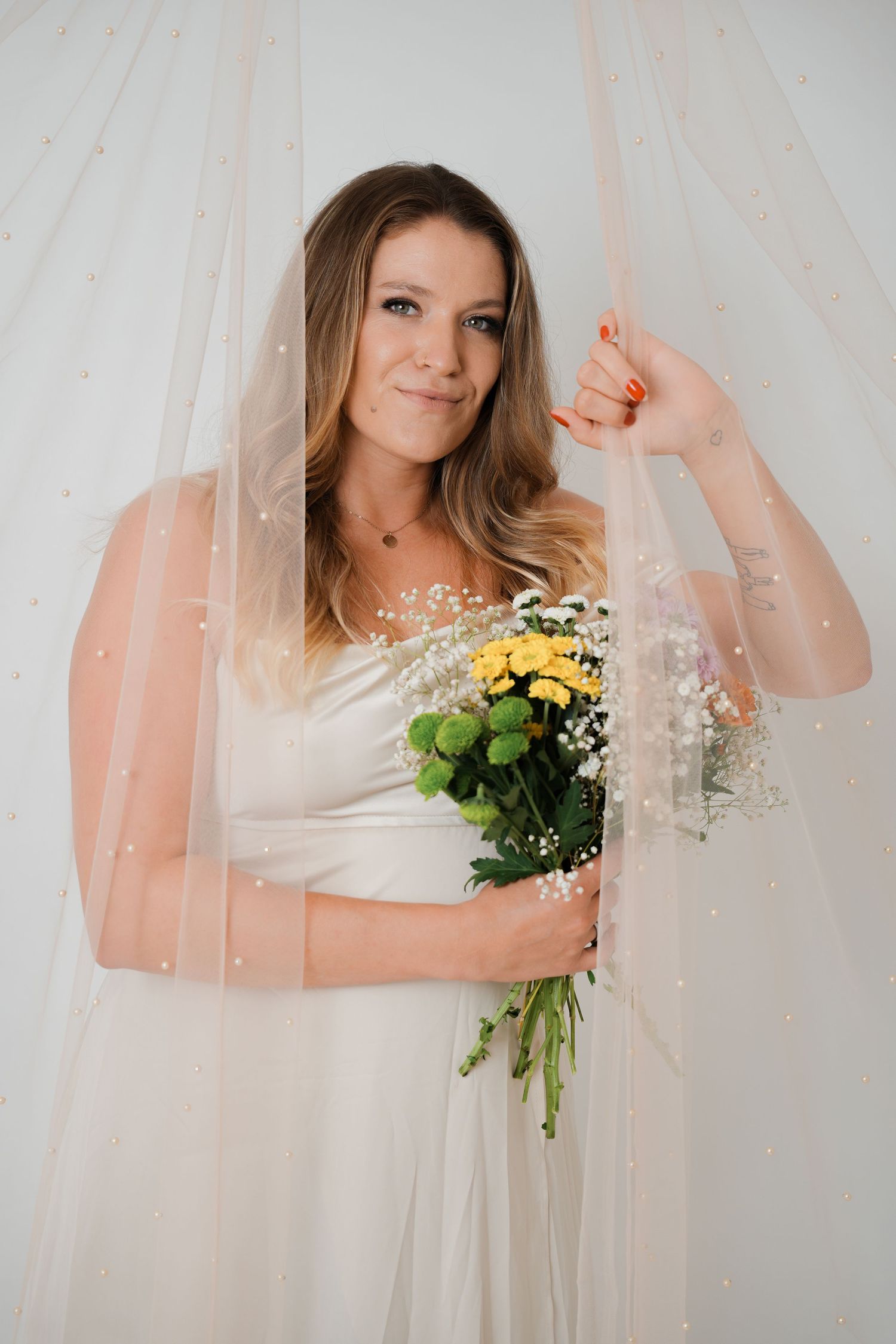 Bride in white gown and pearl veil holds yellow and white flower bouquet, posing elegantly in a soft studio setting.