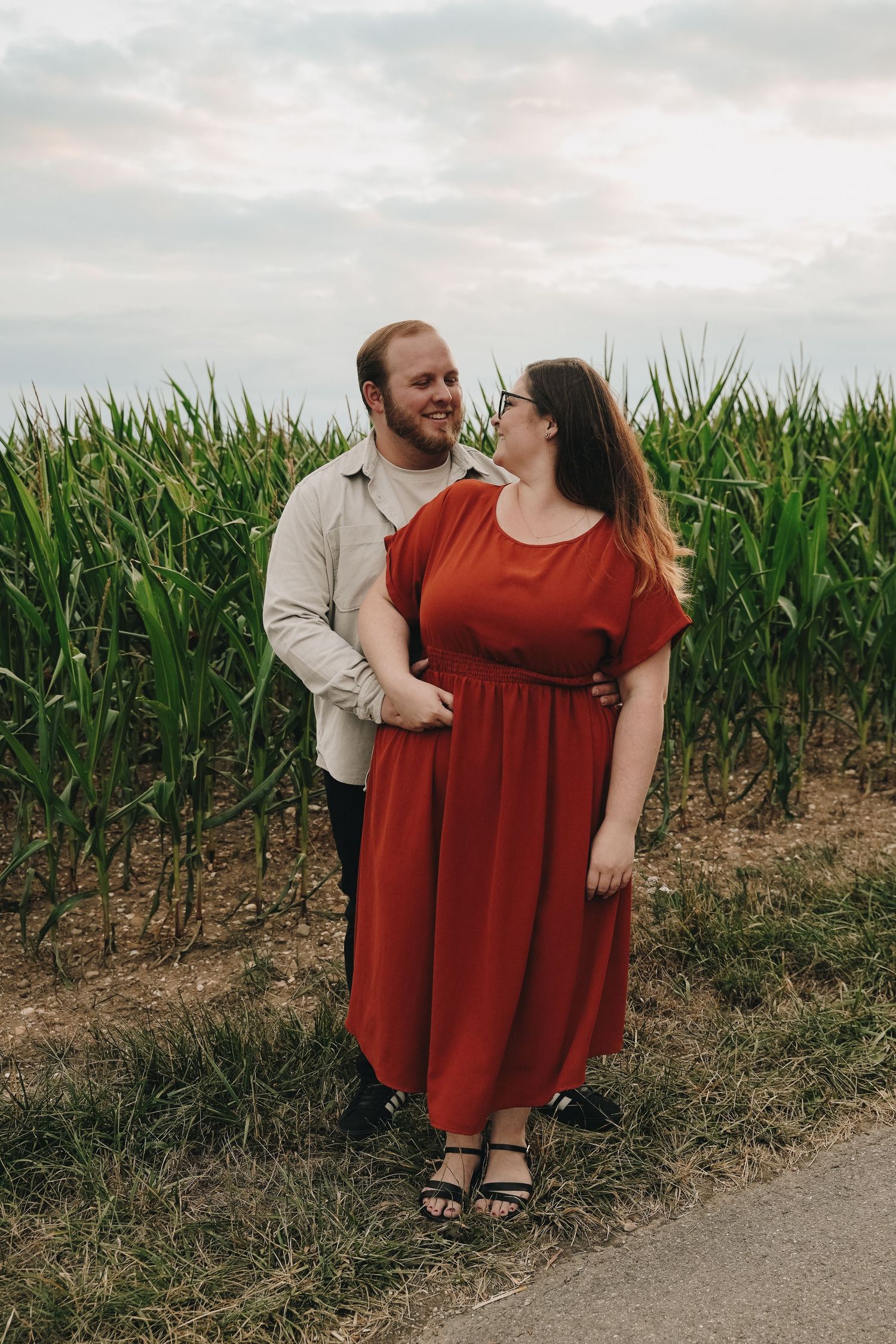 Couple embracing in front of a green cornfield, woman in red dress and man in beige shirt, smiling at each other.