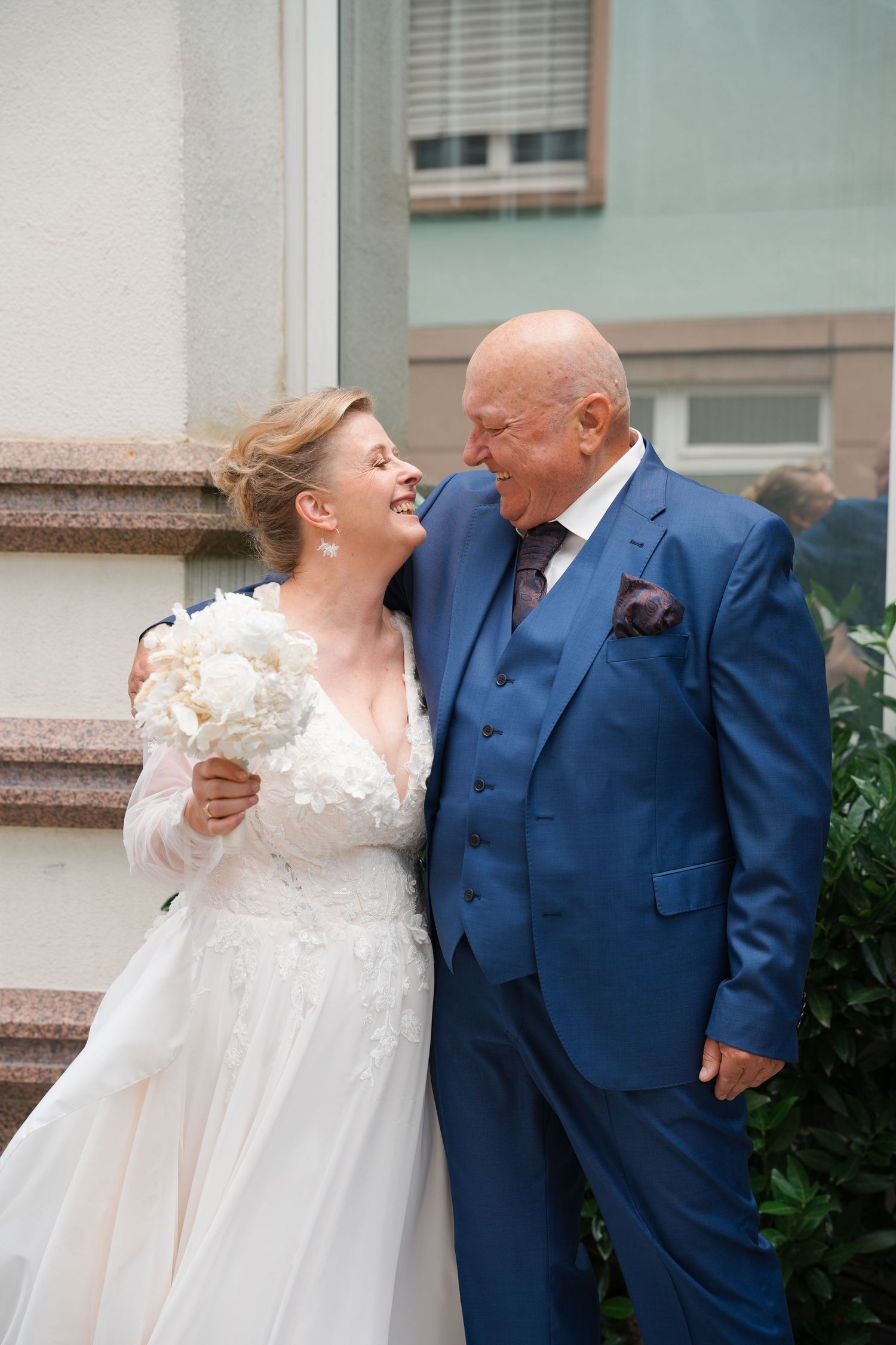 Elderly groom in blue suit and bride in white dress laughing together outdoors on their wedding day.