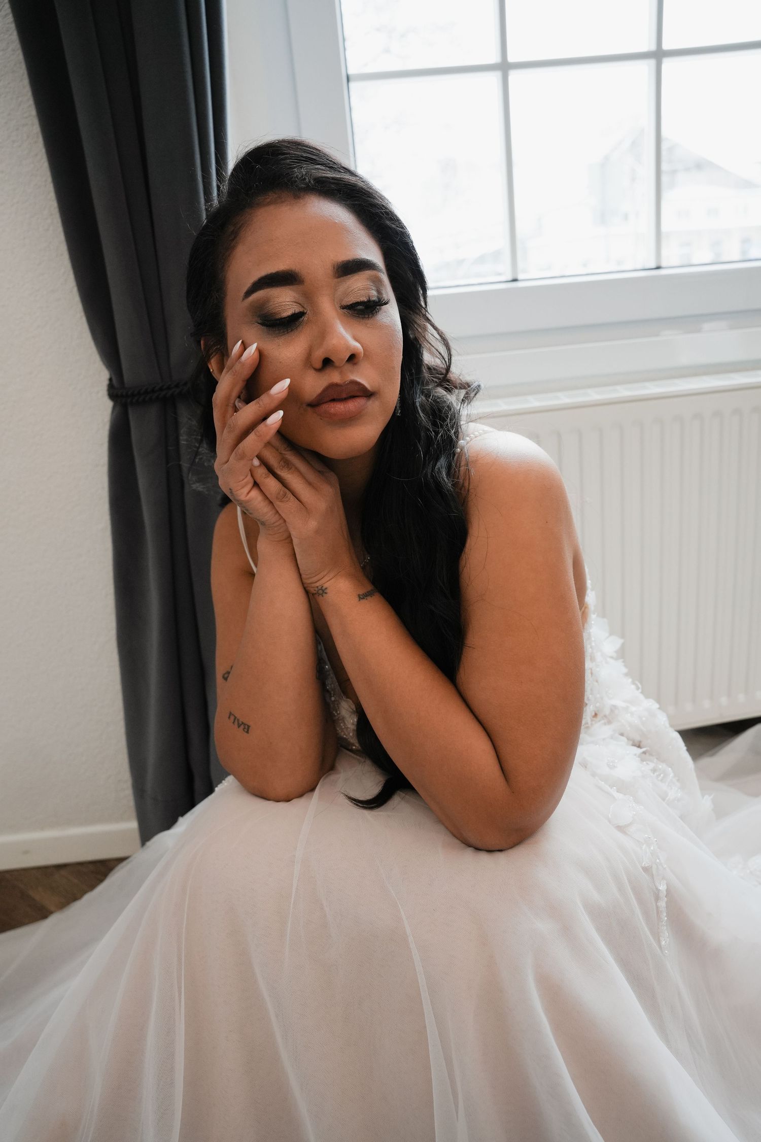 A bride in a white gown sits gracefully by a window, eyes closed, hands folded near her face, wearing elegant makeup.