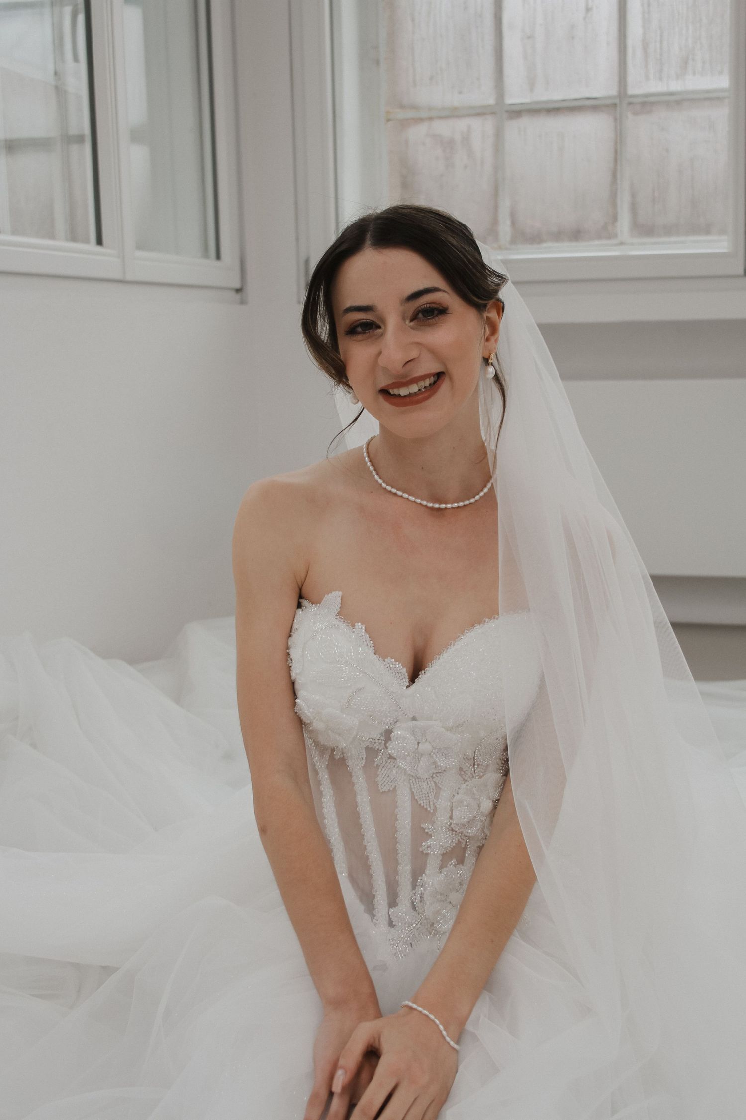 Smiling bride in a strapless lace corset wedding gown with tulle veil and pearl necklace seated by window.