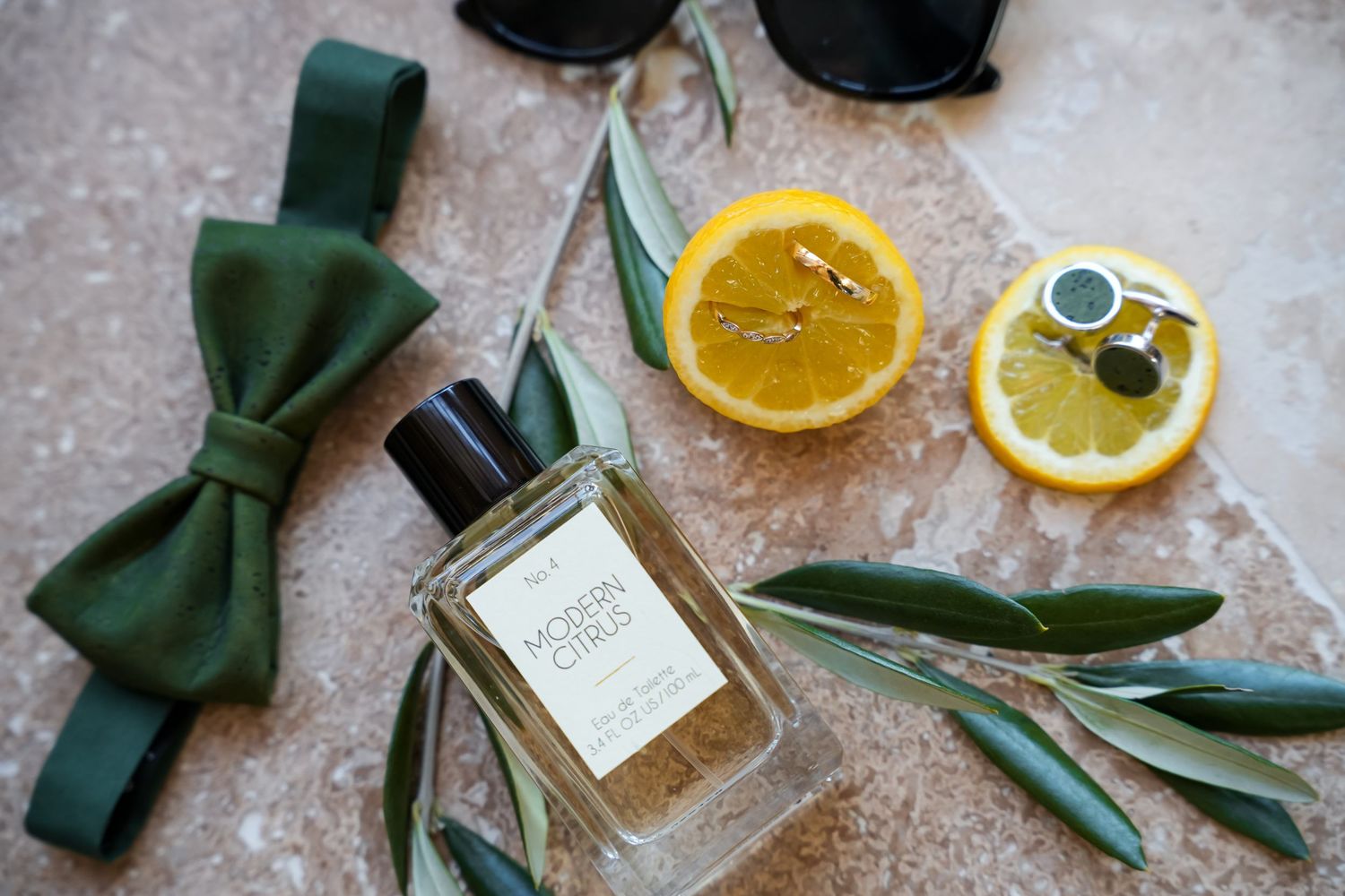 Green bow tie, Modern Citrus cologne, lemon slices, gold cufflinks, and olive branches styled as groom accessories.