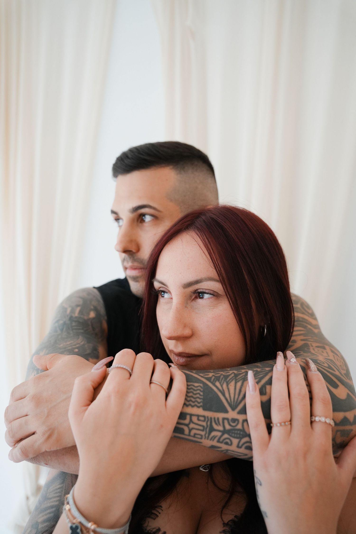 A tattooed couple embracing, woman with red hair in front, man with sleeve tattoos wrapping arms around her near white curtains.