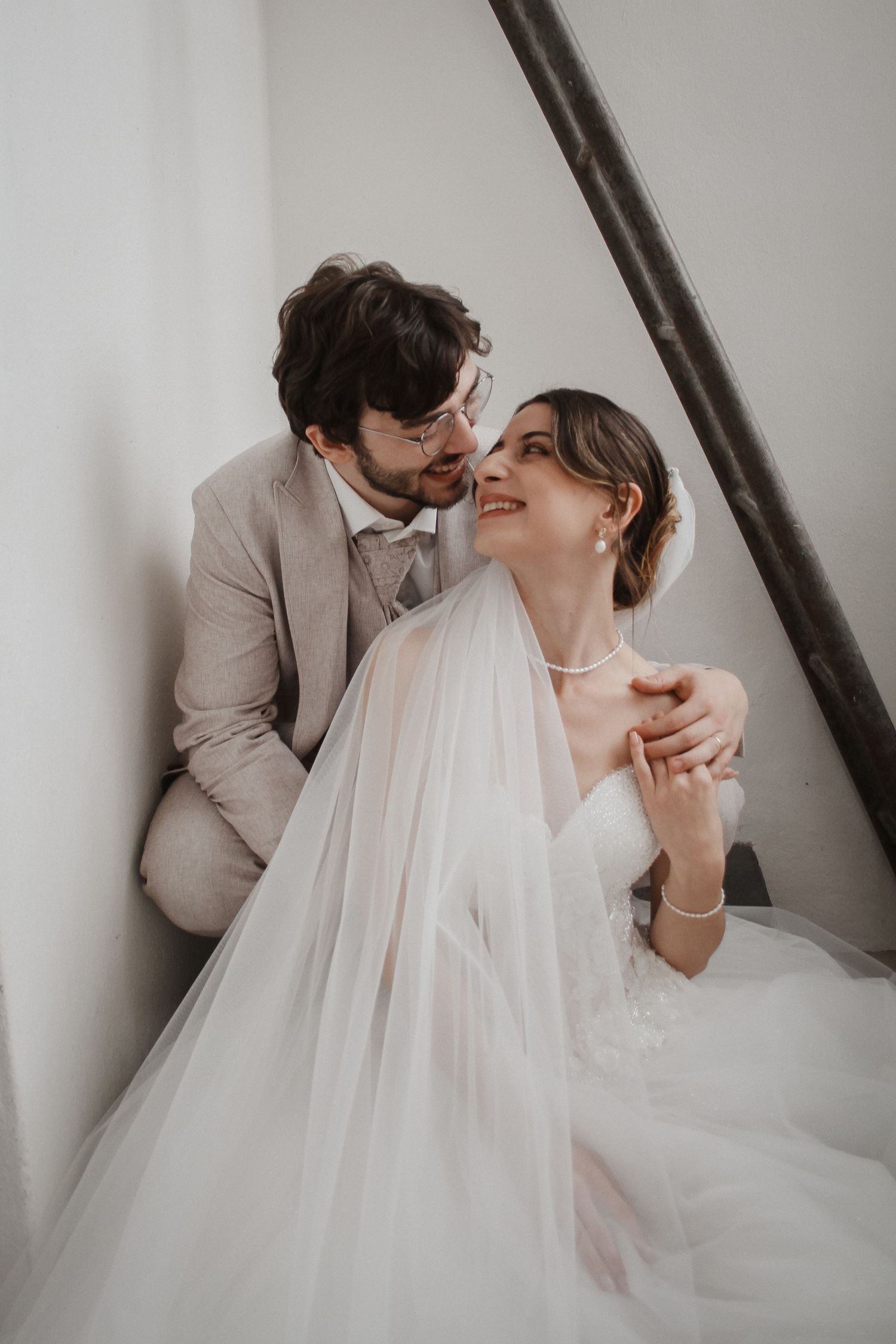 A smiling bride in a white gown and veil sits with a groom in a beige suit, sharing a tender moment together.
