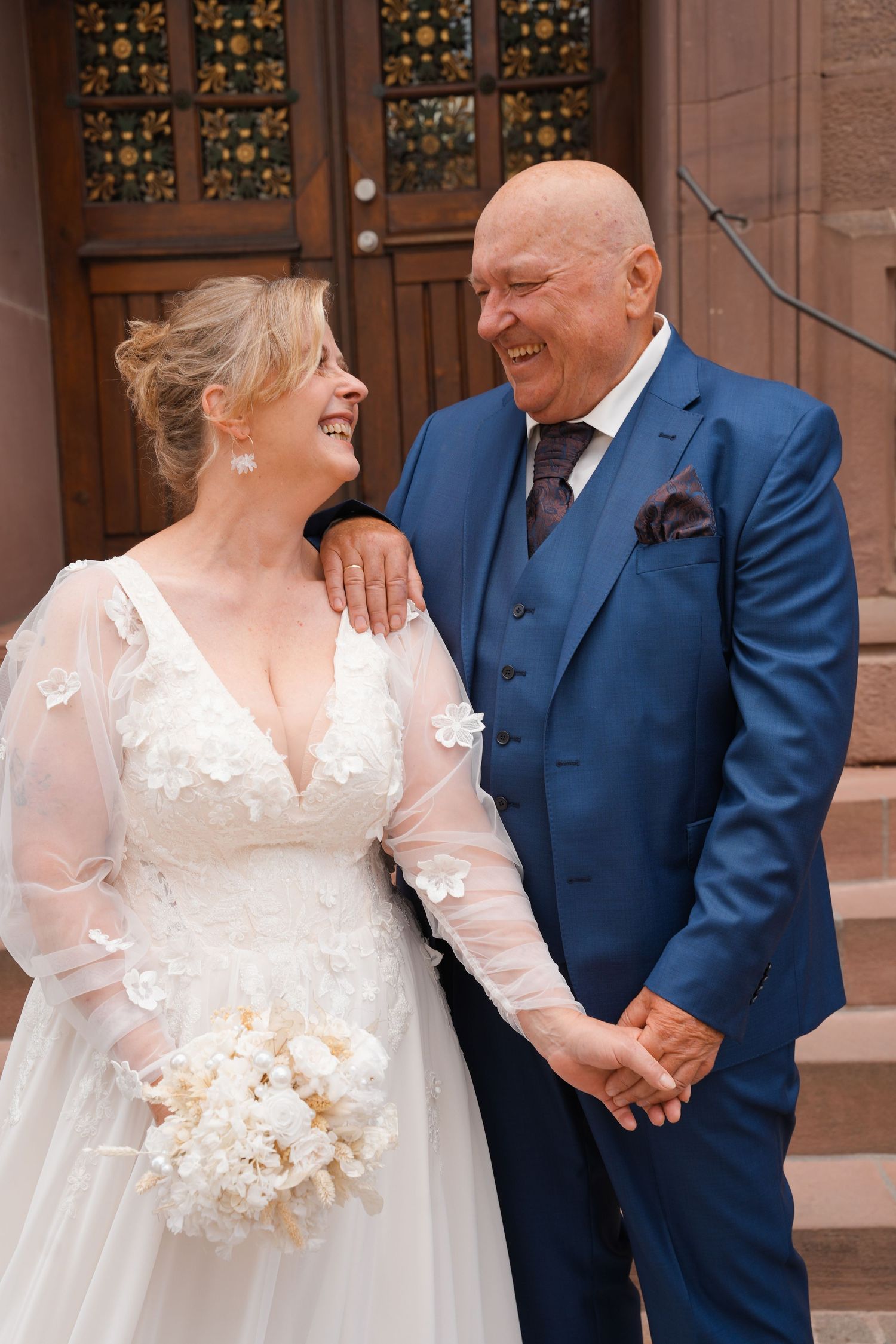 Joyful mature bride in white gown and groom in blue suit laughing together on church steps on their wedding day.