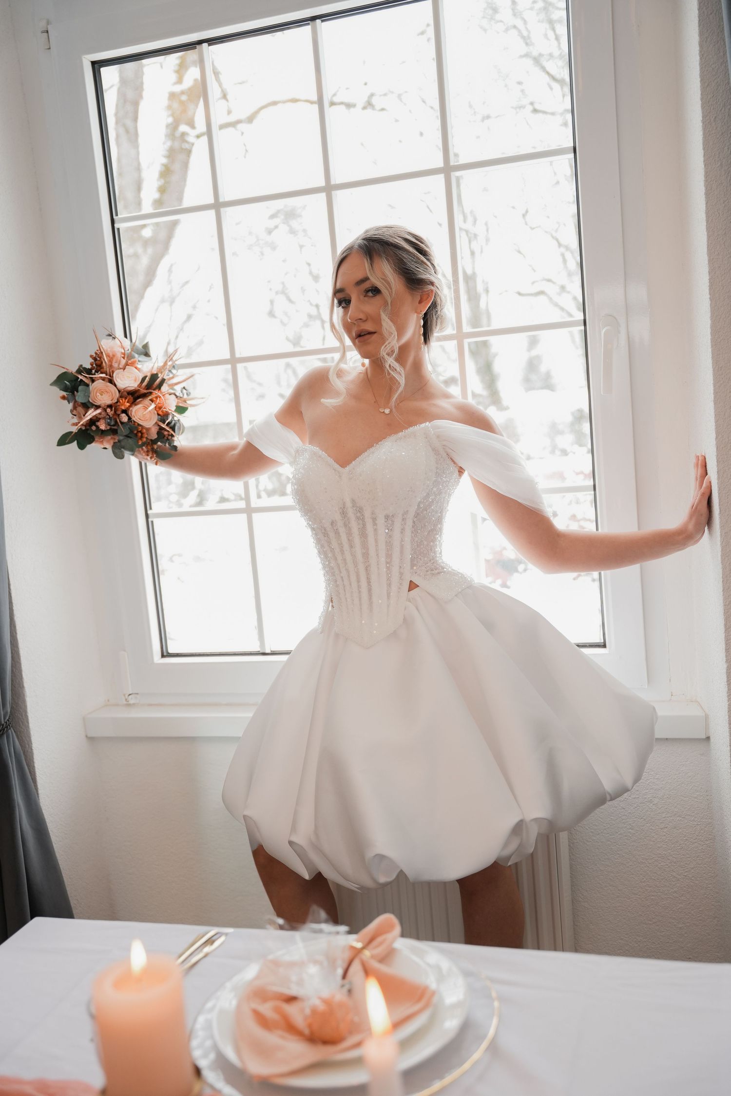Bride in short white off-shoulder wedding dress holding bouquet, posing by bright window with candles on table.