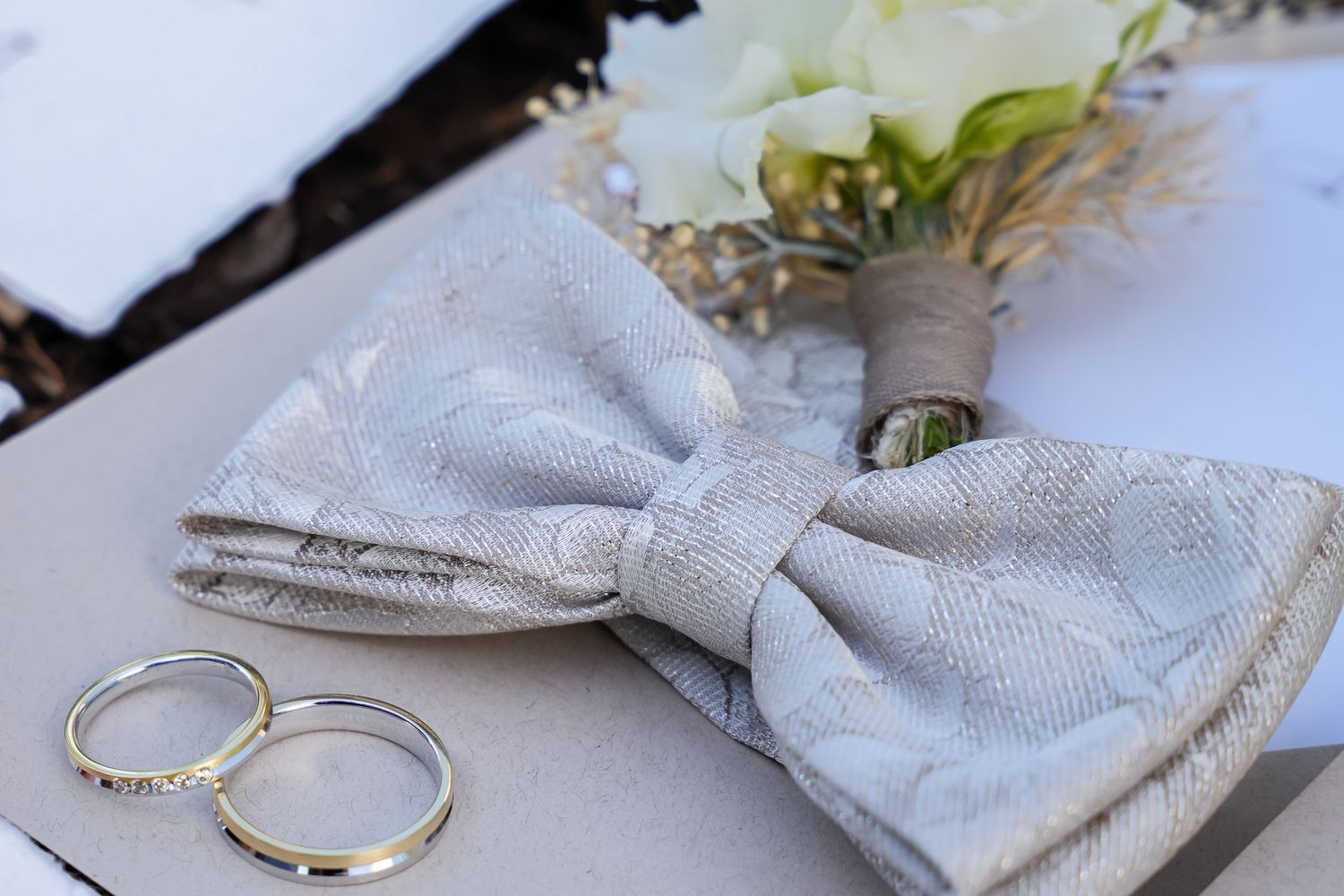 Silver bow tie and wedding rings beside a white floral boutonniere on a light surface.