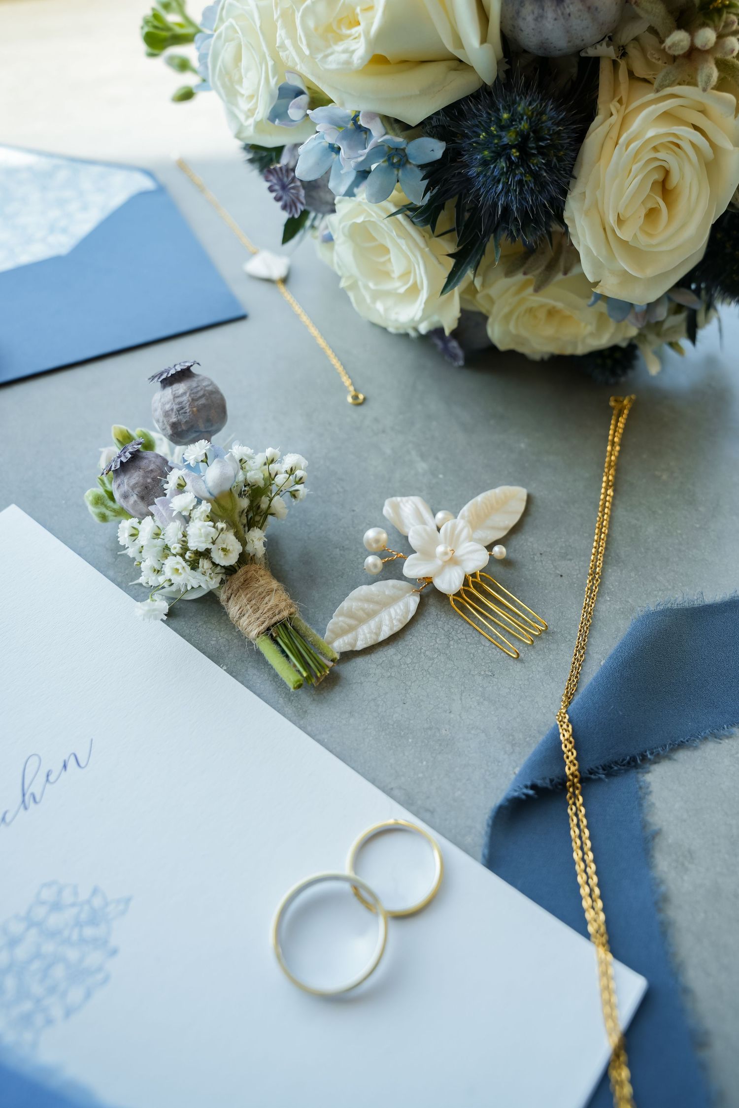 Elegant wedding flat lay with gold rings, floral hair comb, boutonniere, blue envelopes, gold necklace, and white rose bouquet.