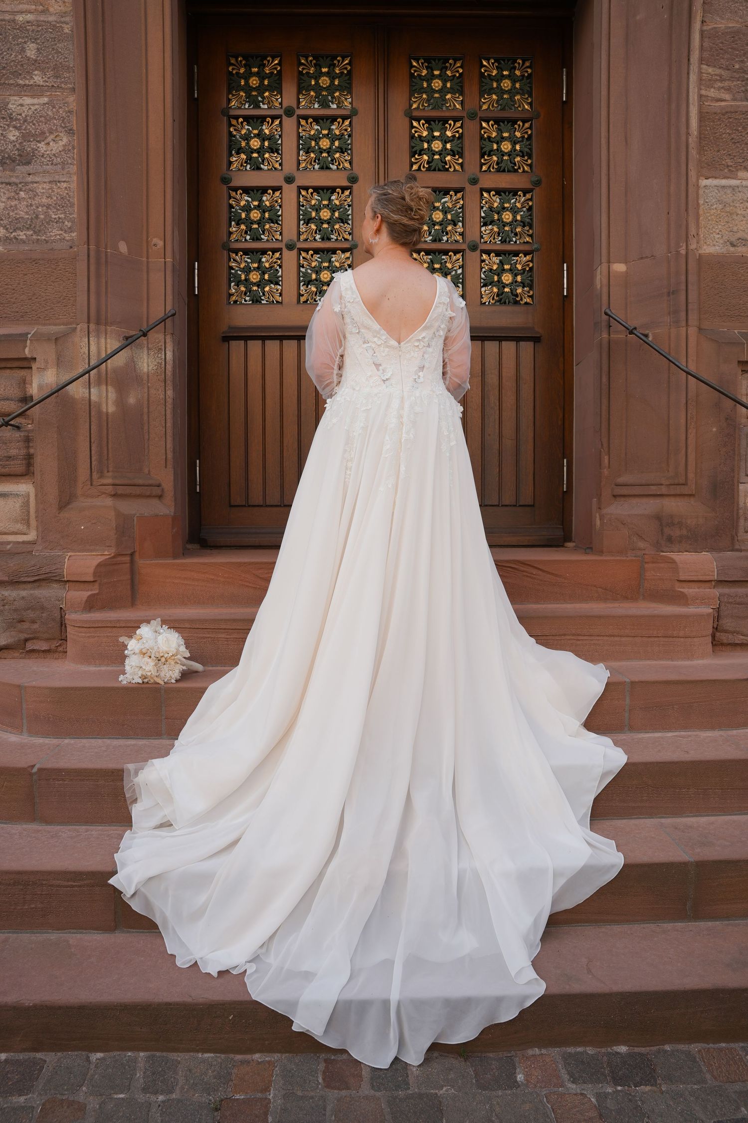 Bride in elegant white gown with lace back and flowing train standing before ornate wooden church doors.