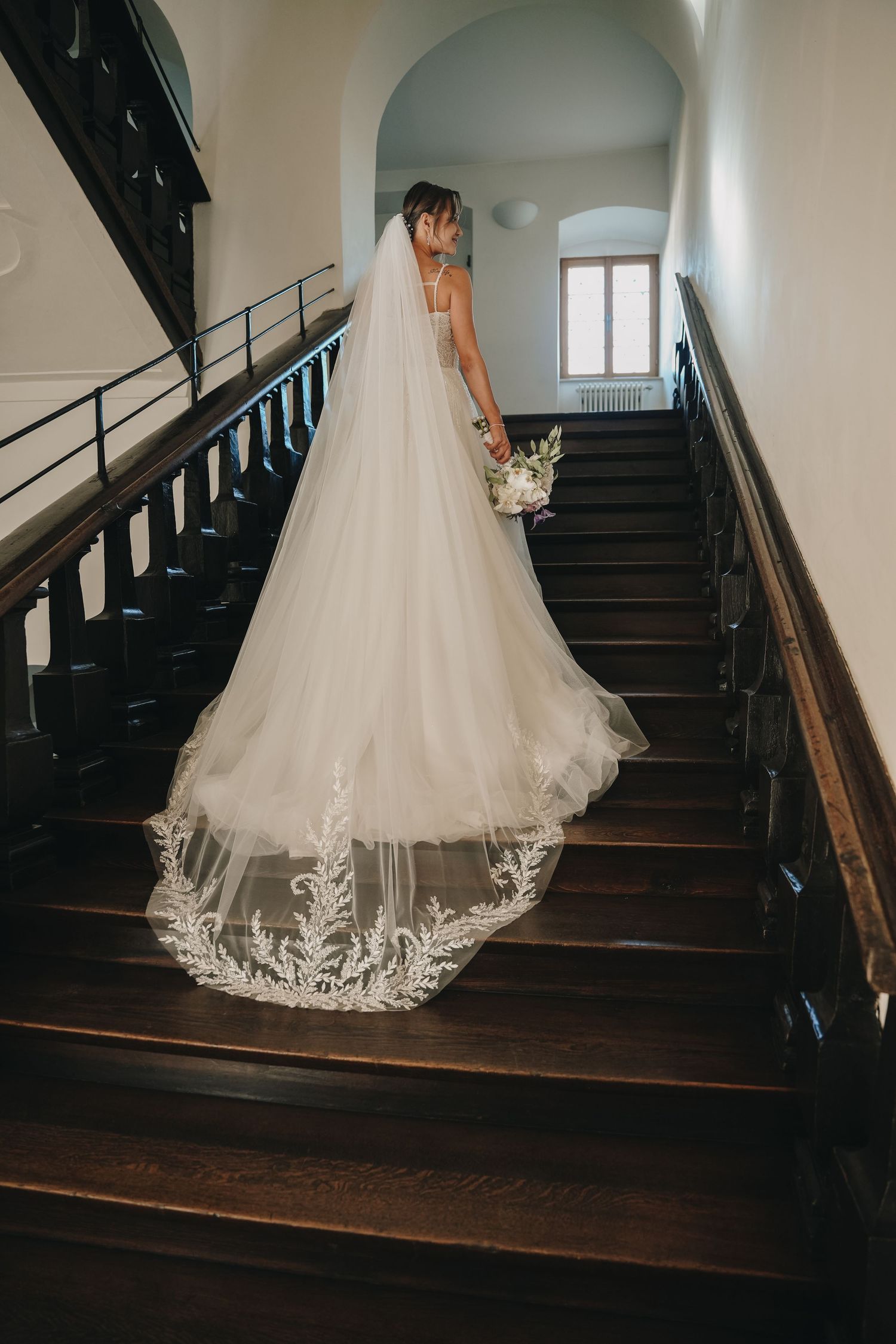 Bride in elegant white gown with lace-trimmed cathedral veil descending dark wooden staircase holding bouquet.