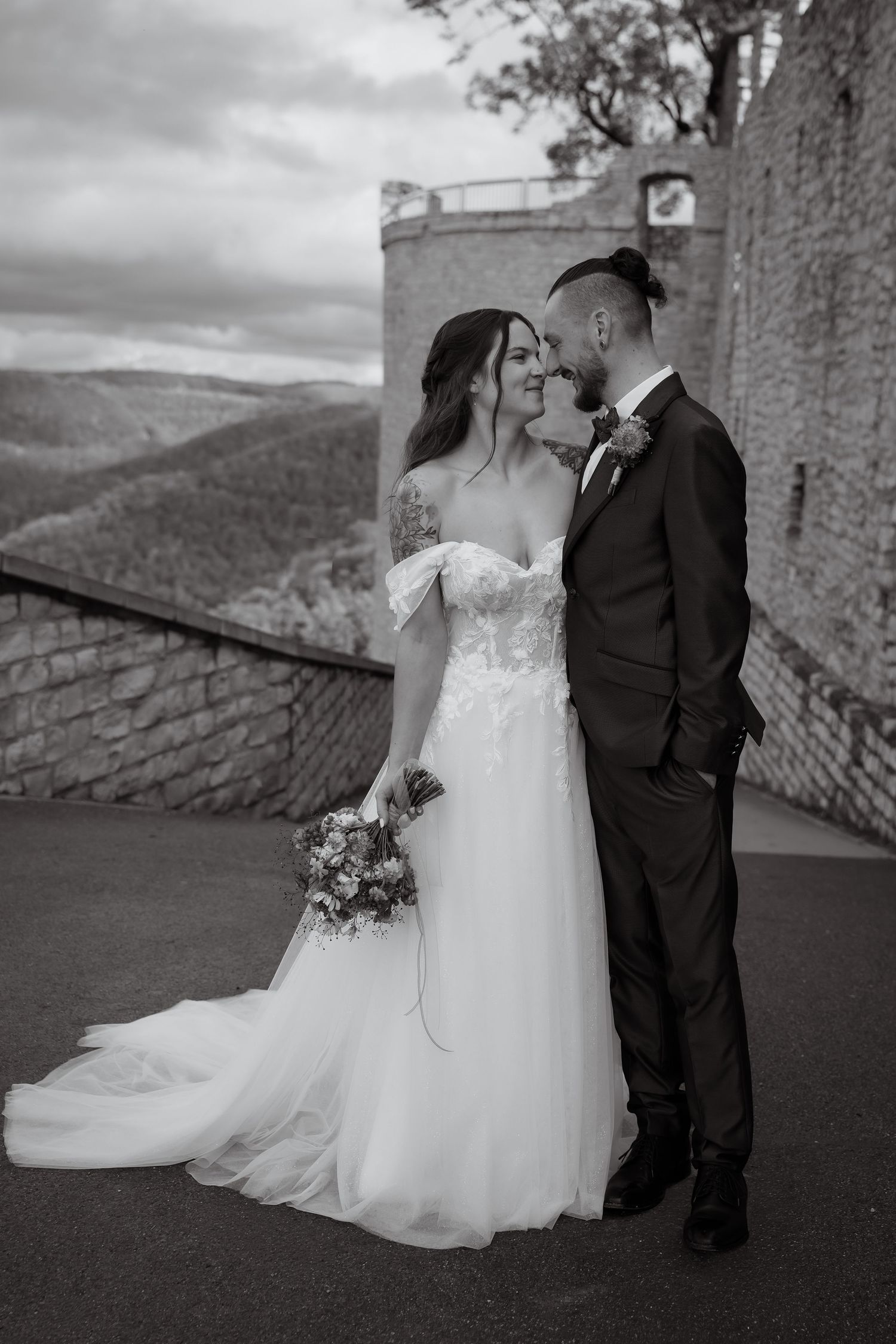 Black and white wedding photo of a bride in an off-shoulder lace gown and a groom in a suit embracing near a stone wall with hills behind.