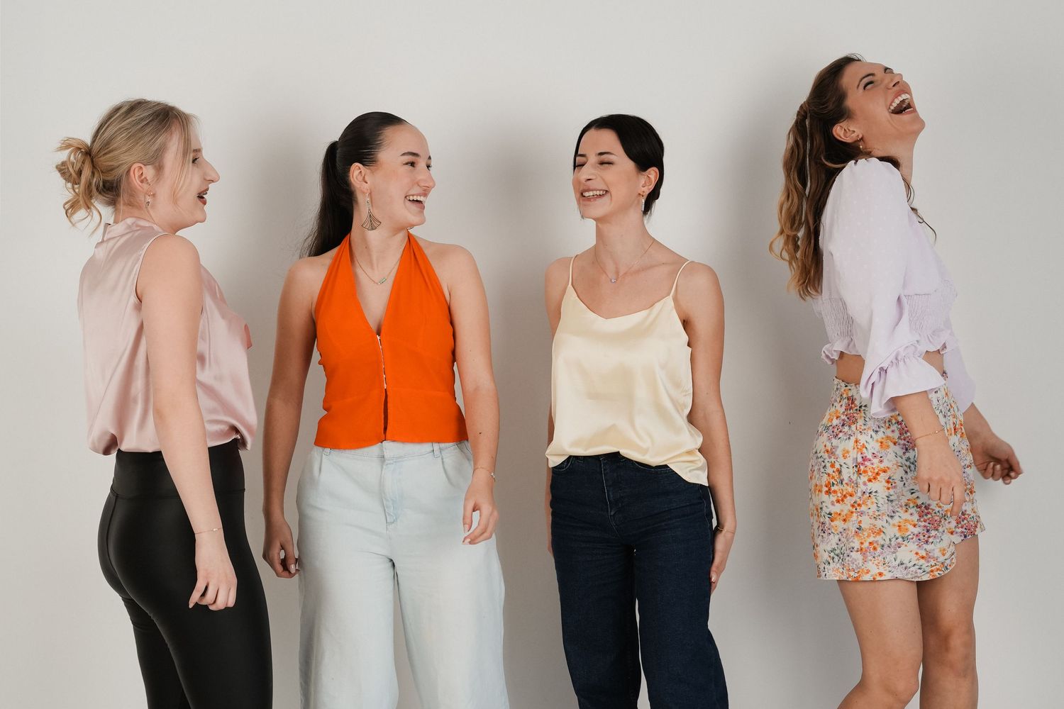 Four stylish women standing together laughing and posing in casual summer outfits against a white background.